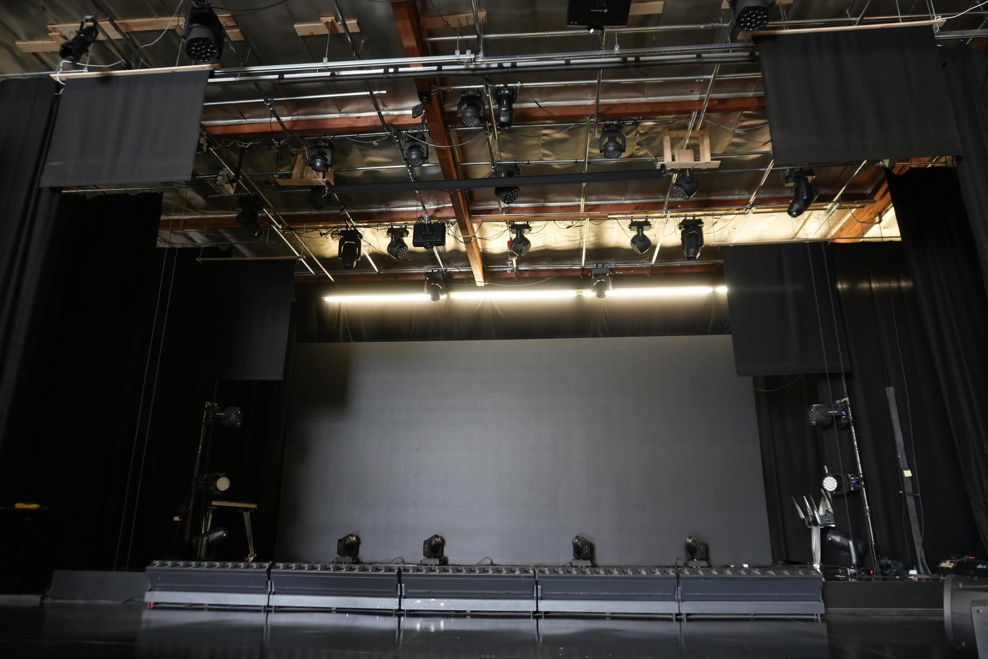 The image shows an empty stage with lighting fixtures mounted above and a large black backdrop.