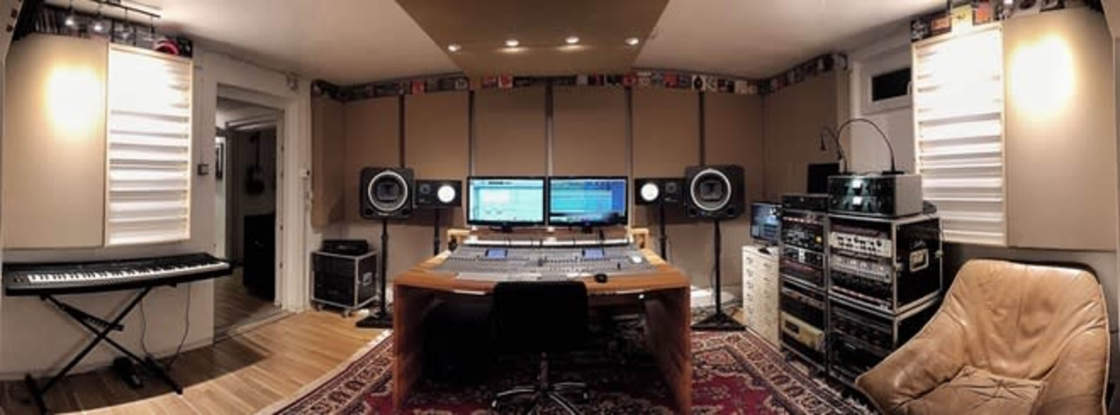 The image shows a music studio featuring a large mixing desk, two computer monitors, speakers, a keyboard, and various audio equipment in a well-furnished setting.