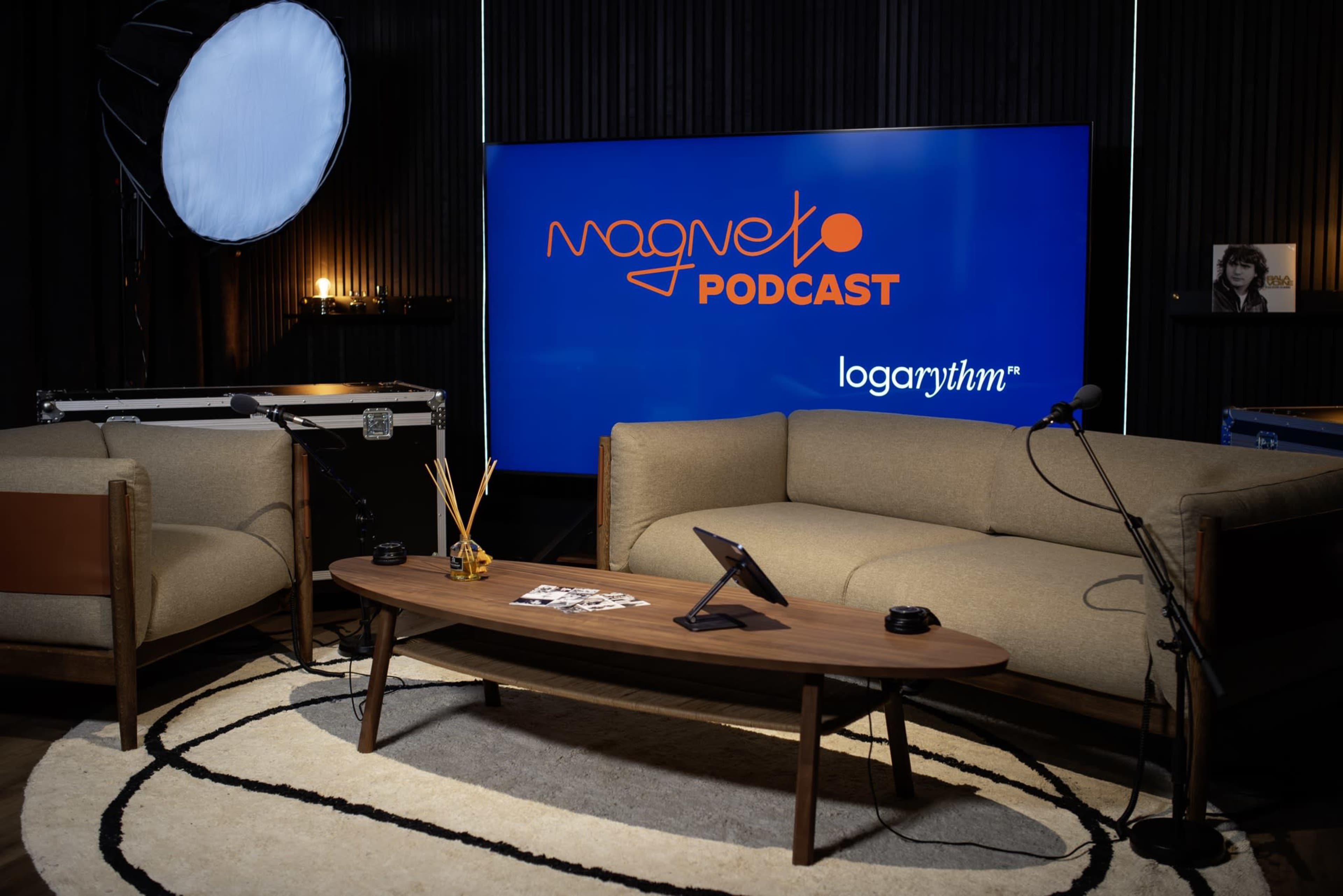 The scene features a modern podcast studio with a large screen displaying the words "Magneto Podcast," two brown couches, and a circular coffee table with decorative items.