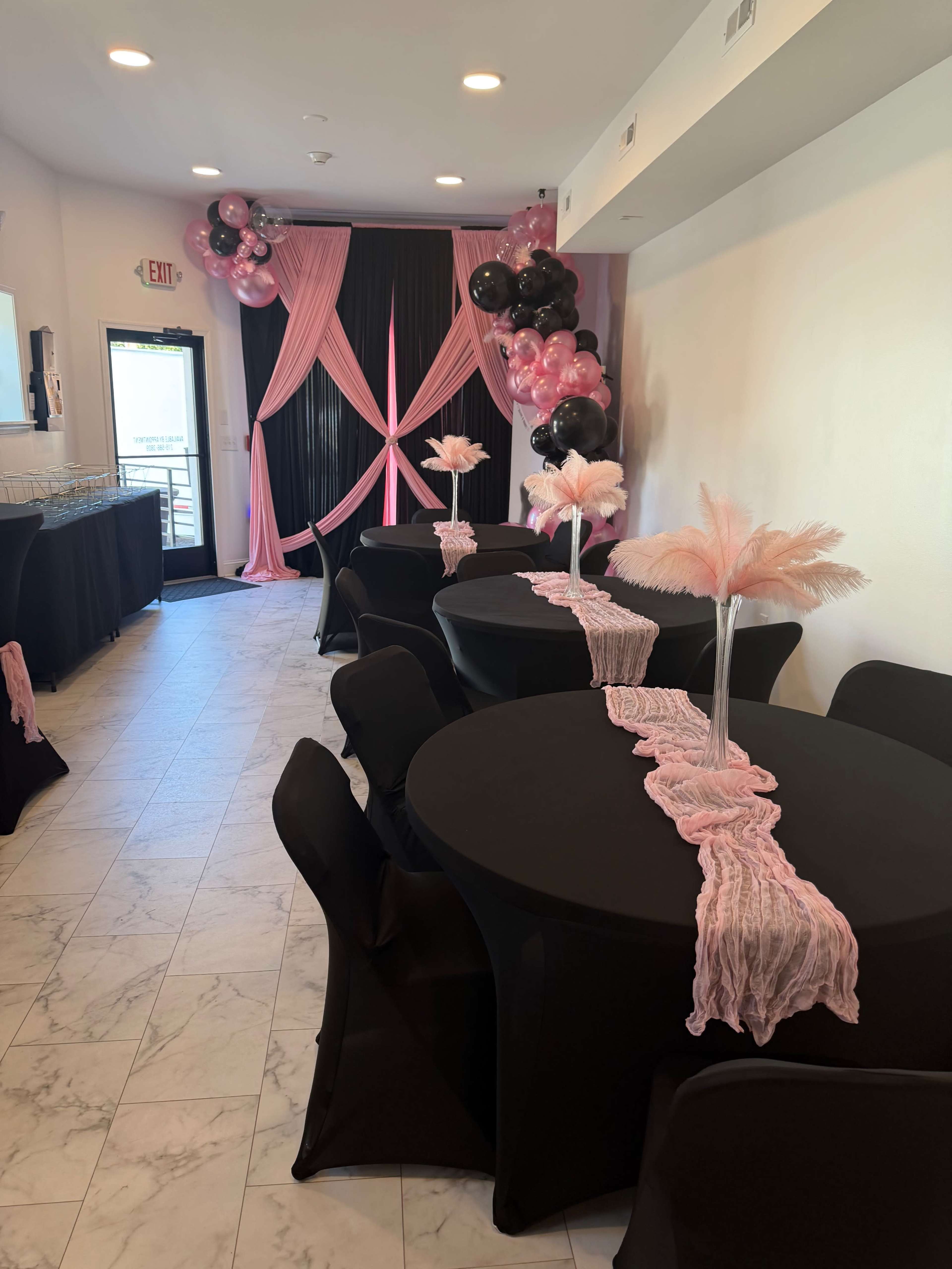 The image depicts a decorated event space with black tables covered in pink runners, surrounded by pink and black balloon arrangements and curtains.
