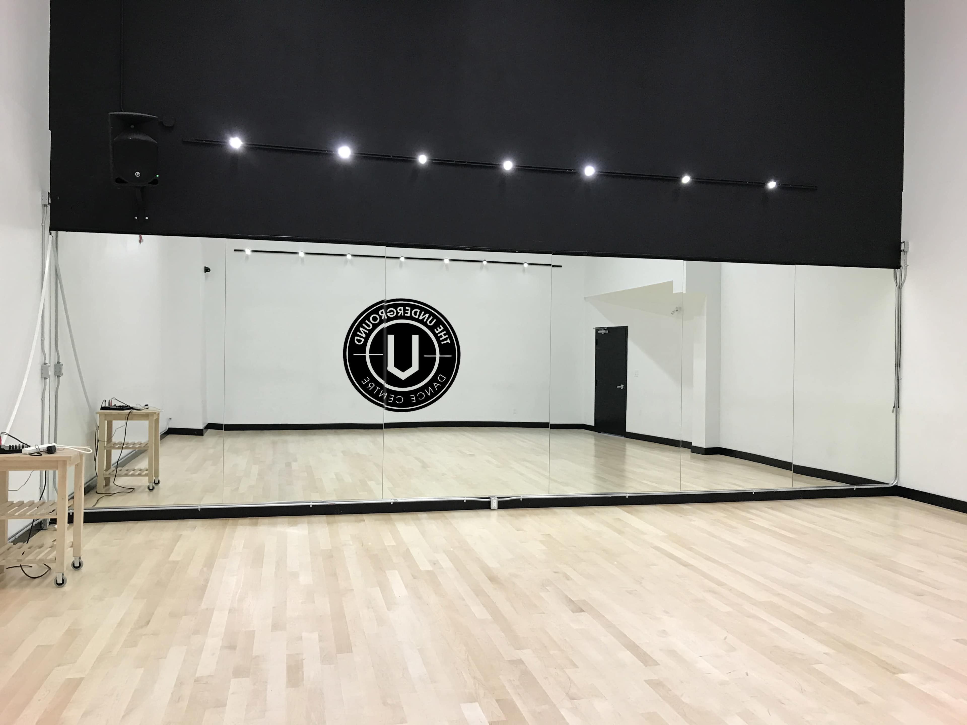 The image shows a spacious dance studio with a large mirrored wall and a logo on the back wall.