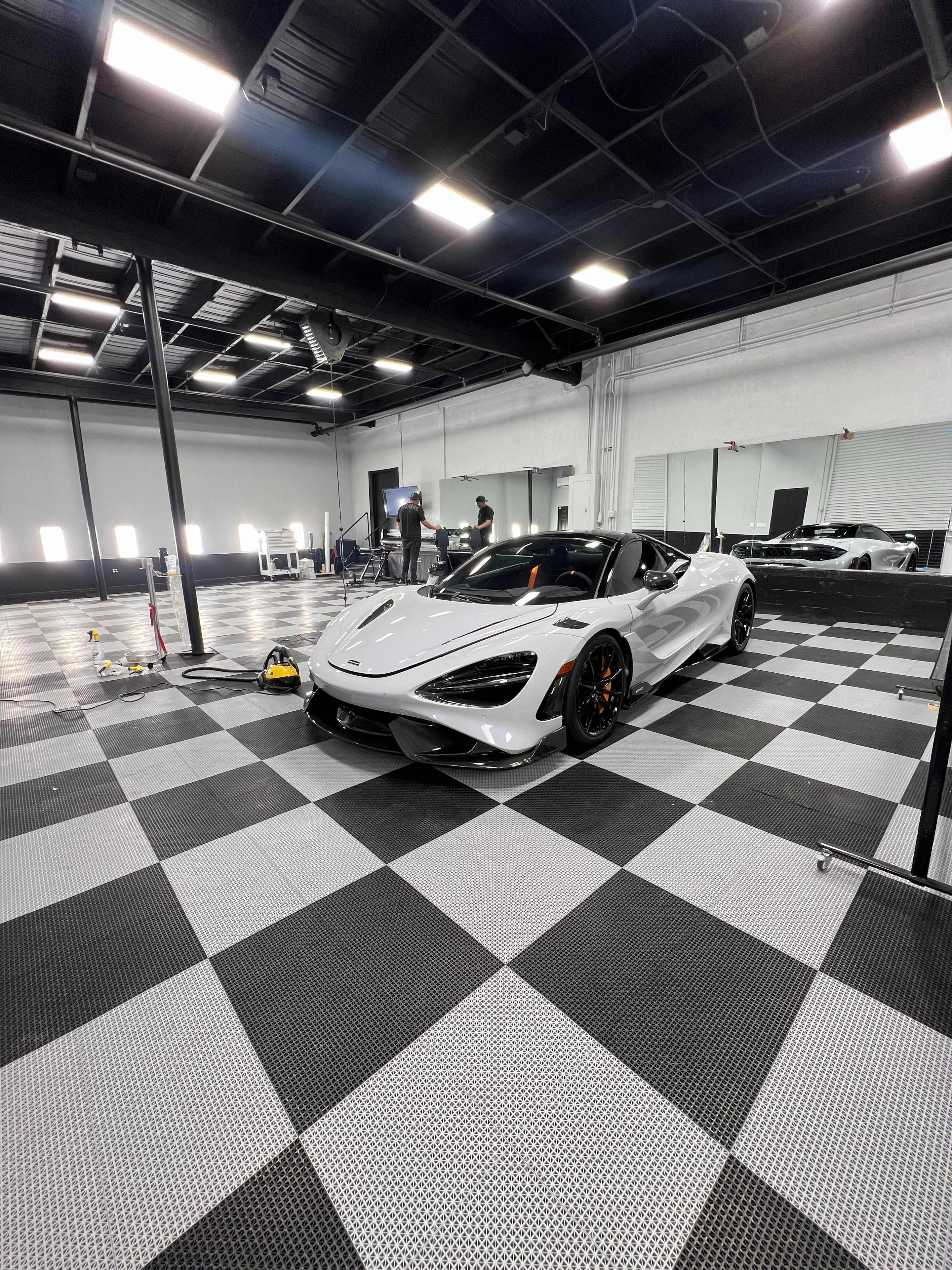 A white McLaren 720S is parked on a checkered floor in a brightly lit garage with several people working in the background.