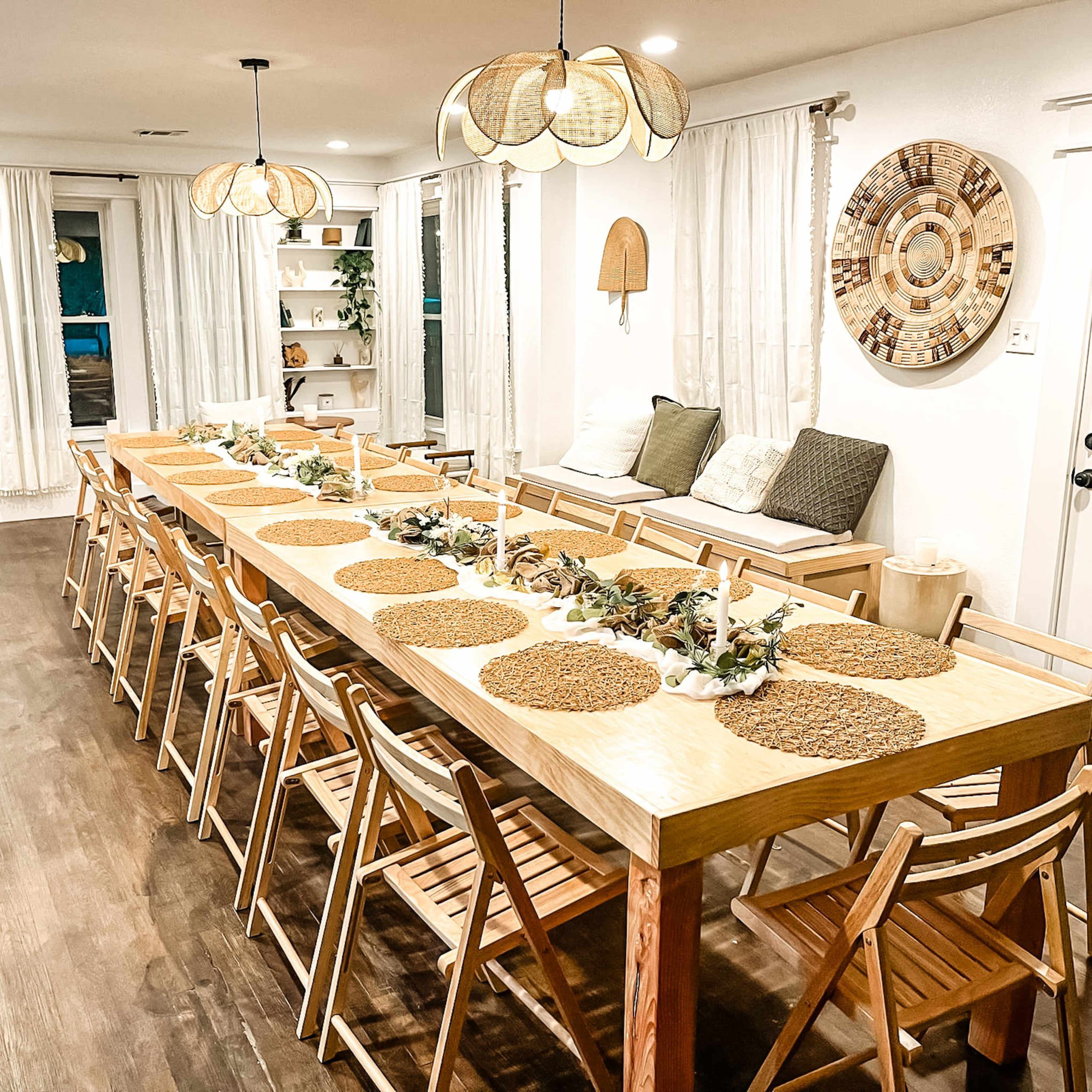 A large wooden dining table is arranged with placemats and decorative elements, surrounded by multiple folding chairs in a well-lit room.
