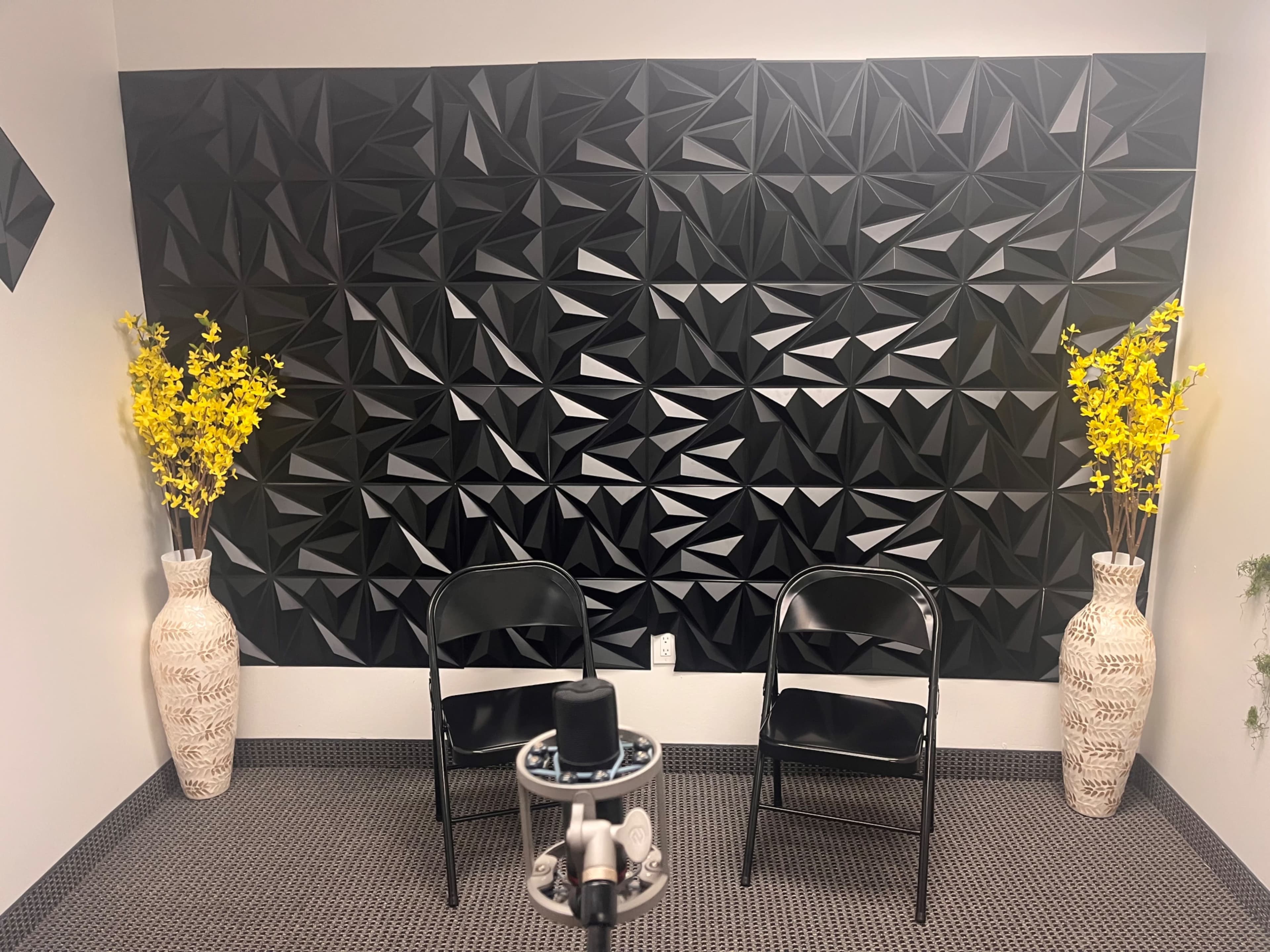 The image shows a small room with a black geometric accent wall, two black folding chairs facing each other, and decorative vases containing yellow flowers on either side.
