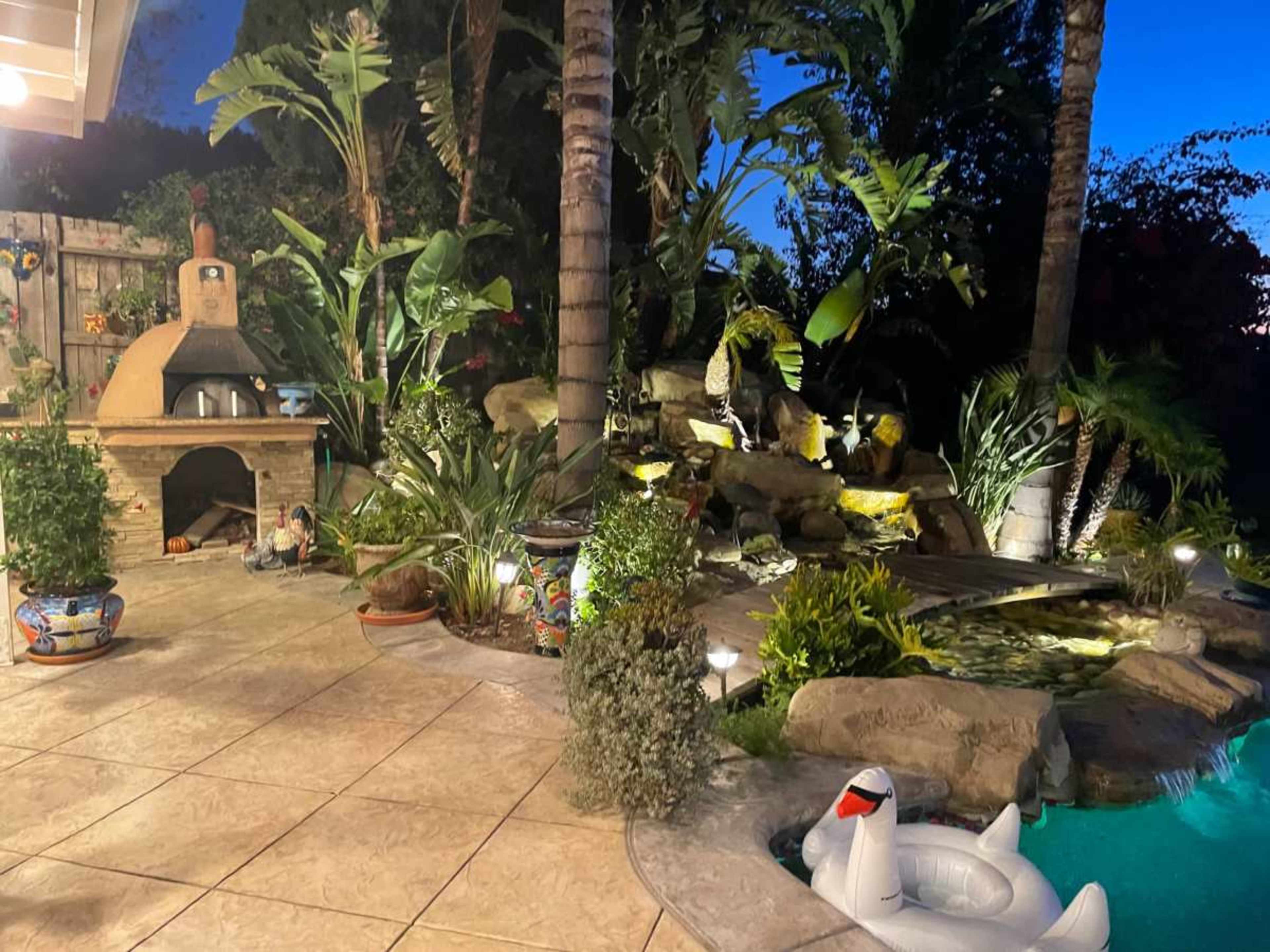 The image shows a well-lit backyard with a stone outdoor fireplace, lush tropical plants, and a swimming pool featuring a large inflatable swan.
