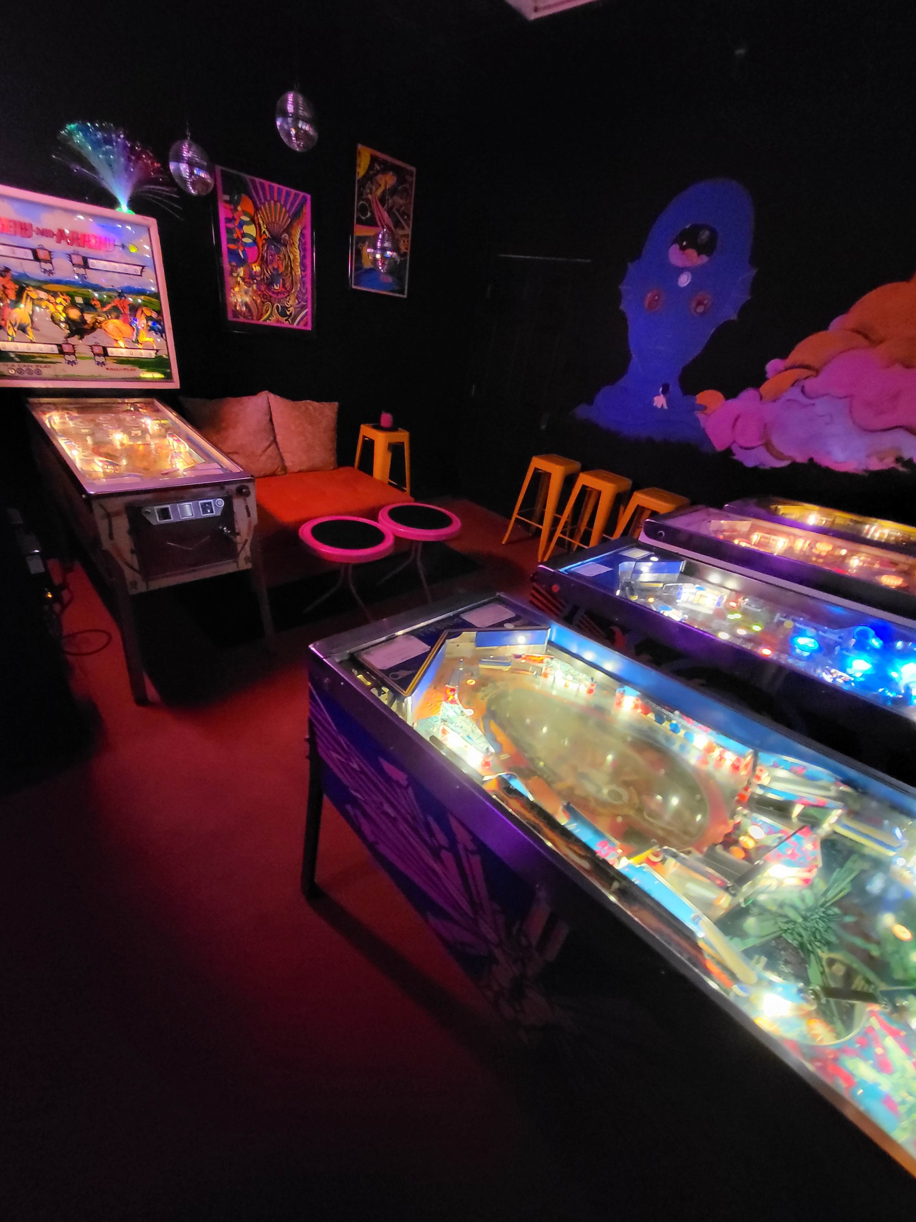 A dimly lit room filled with illuminated pinball machines, colorful artwork on the walls, and seating arrangements in the background.