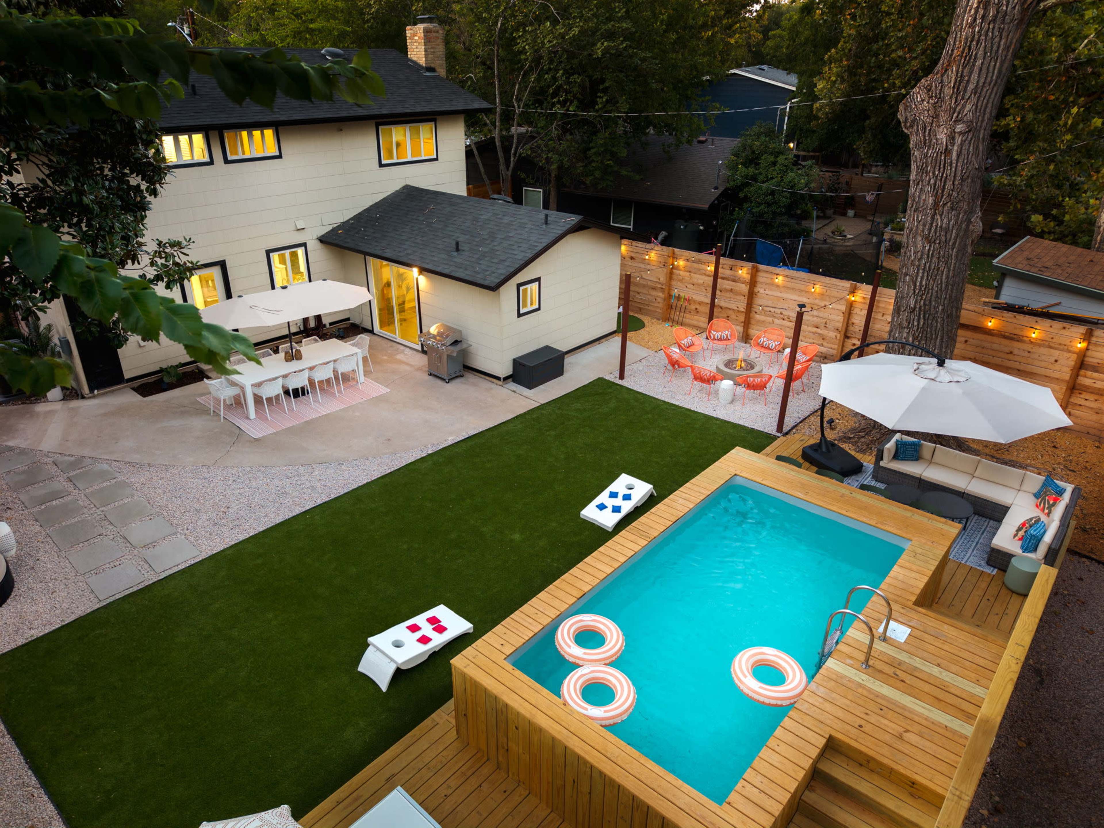The image shows a backyard scene featuring a wooden deck with a swimming pool, surrounded by green grass, outdoor seating, and a house in the background.