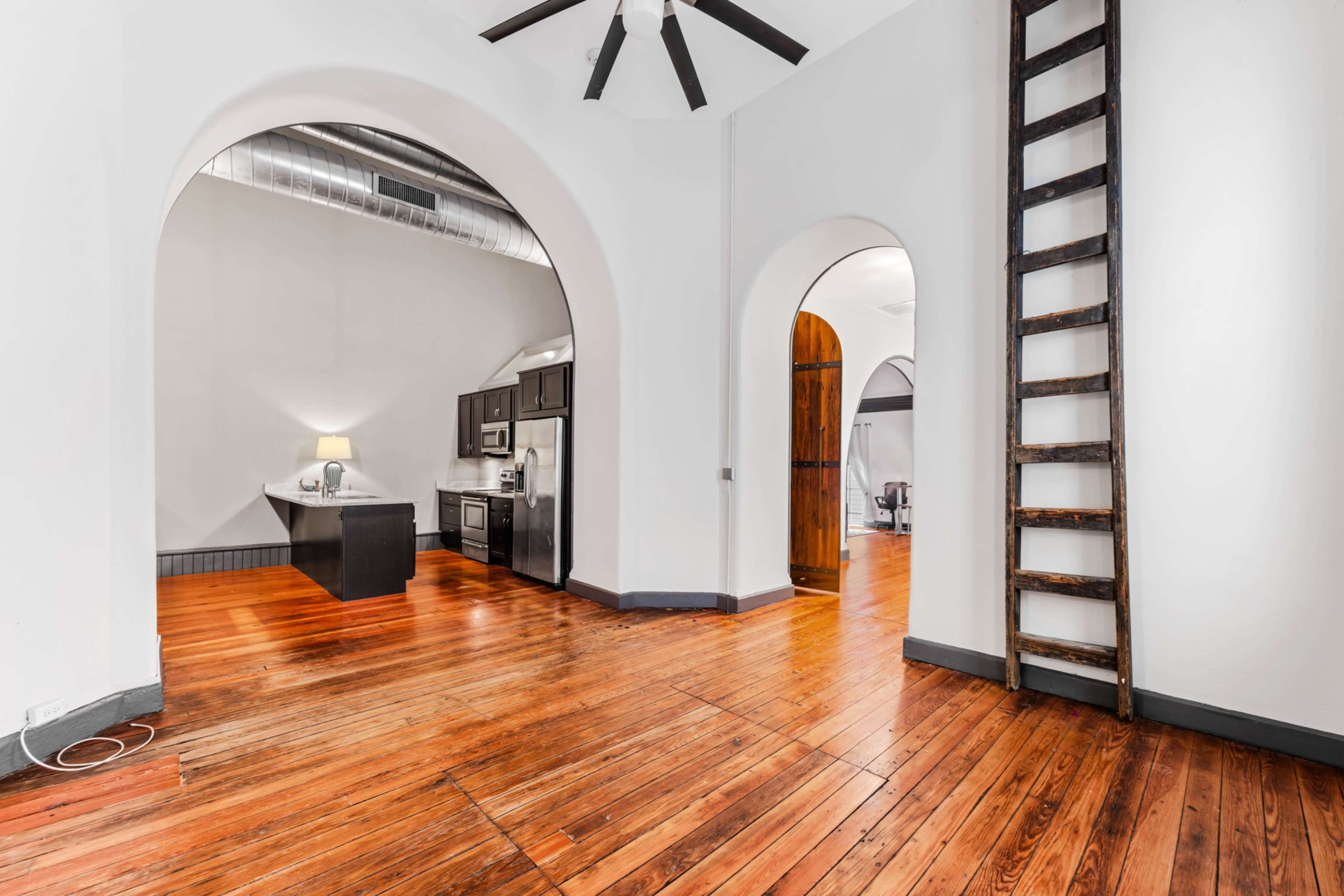 The image shows a spacious room with wooden flooring, a kitchen area featuring stainless steel appliances, and an adjoining ladder against the wall.