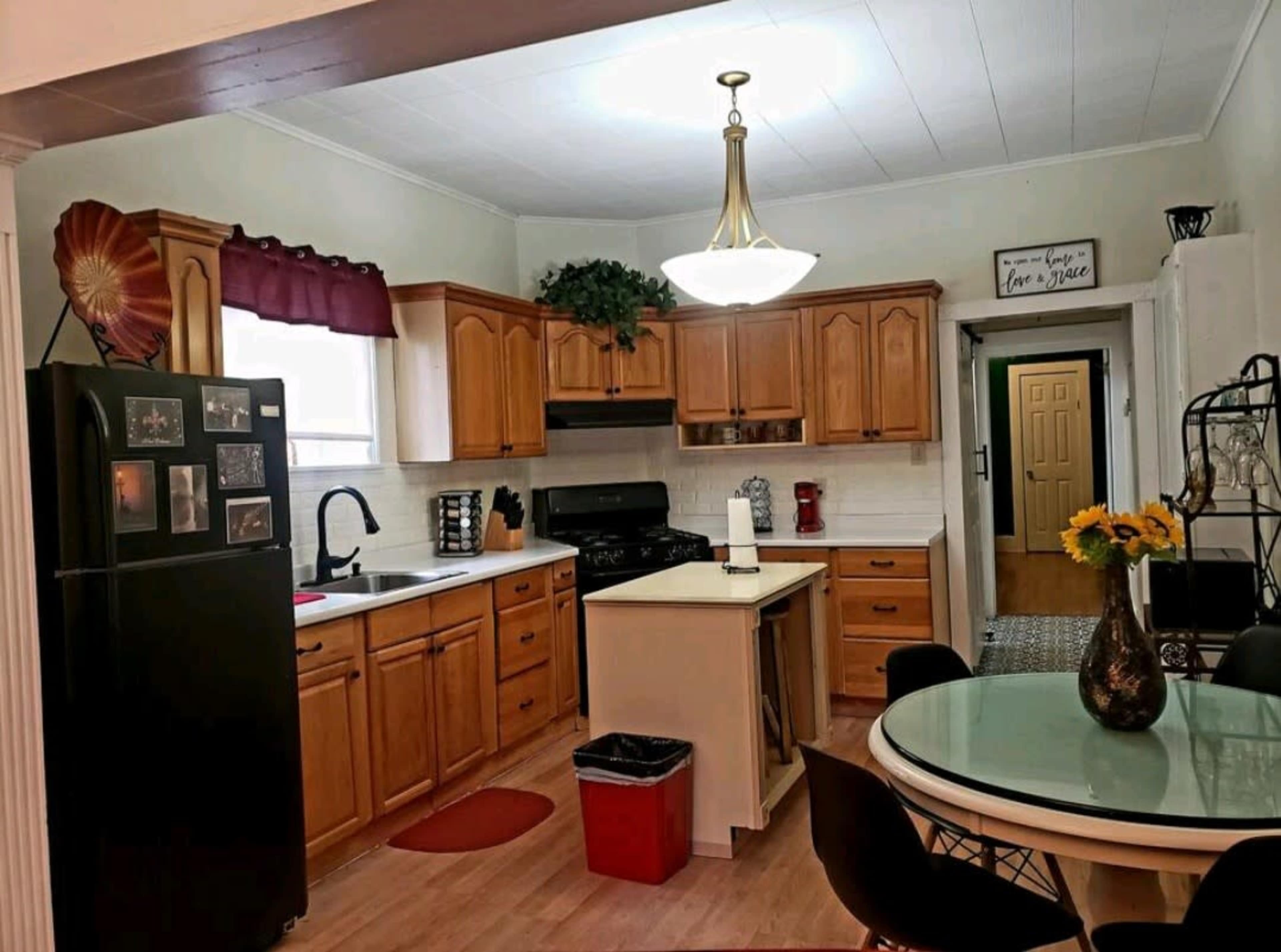 The image shows a kitchen featuring wooden cabinets, a central island, a black refrigerator, and a dining table with chairs.