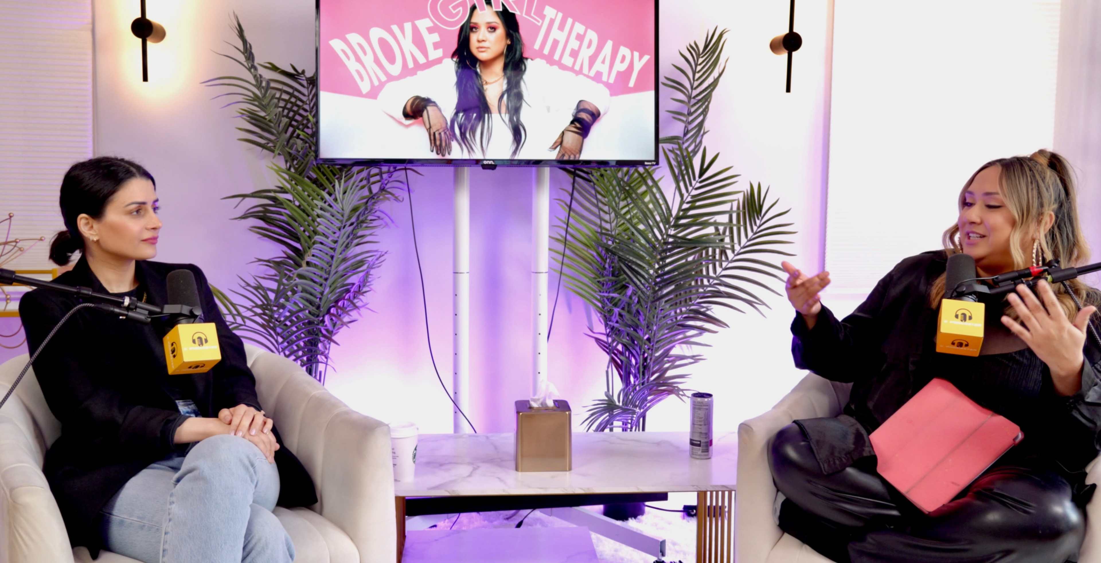 Two women sit in armchairs, speaking across a coffee table, with a television screen behind them displaying a show titled "Broke & Therapy" alongside a graphic of a host.