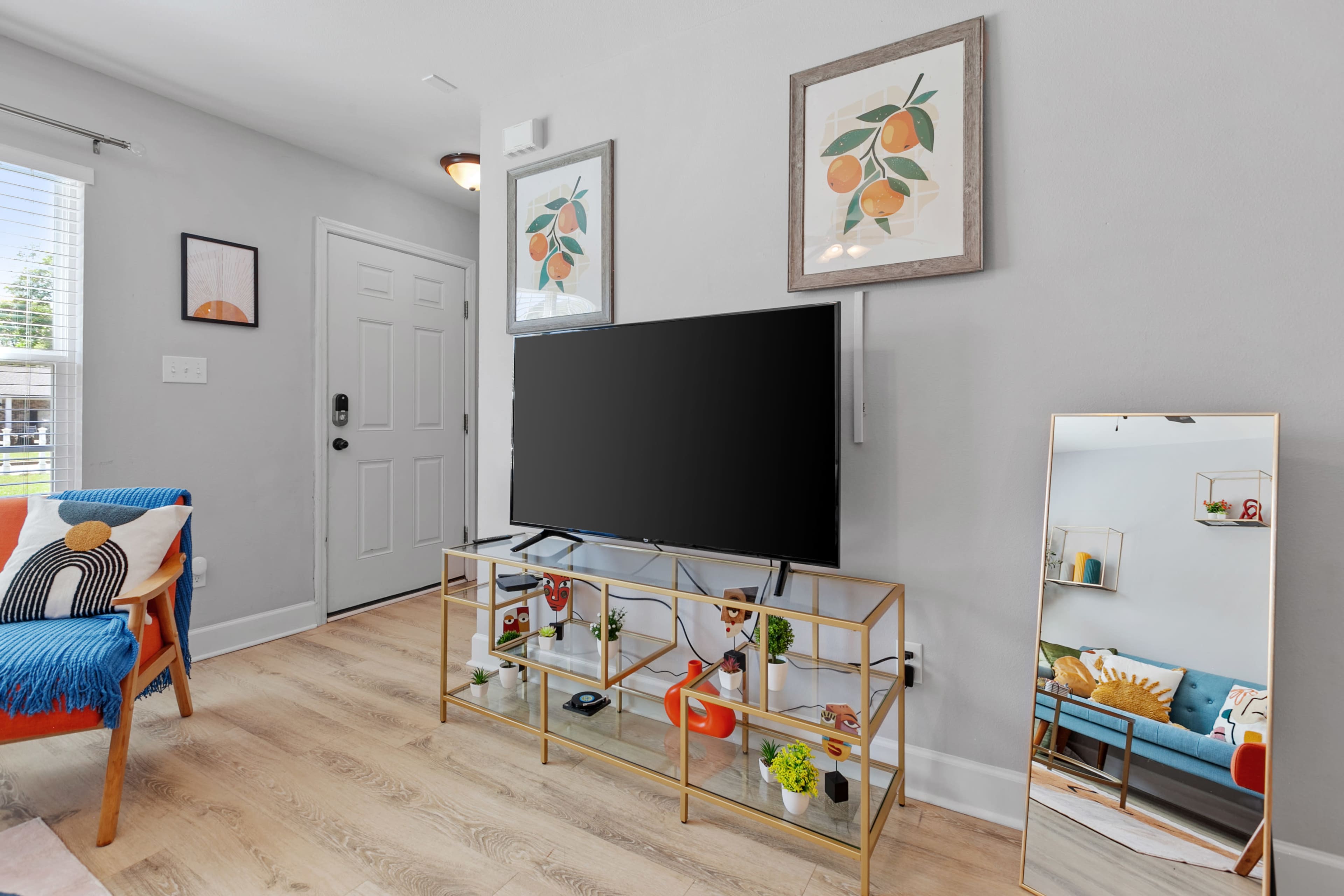 A living room with a television on a glass shelf, framed artwork on the walls, and a full-length mirror beside a colorful chair.