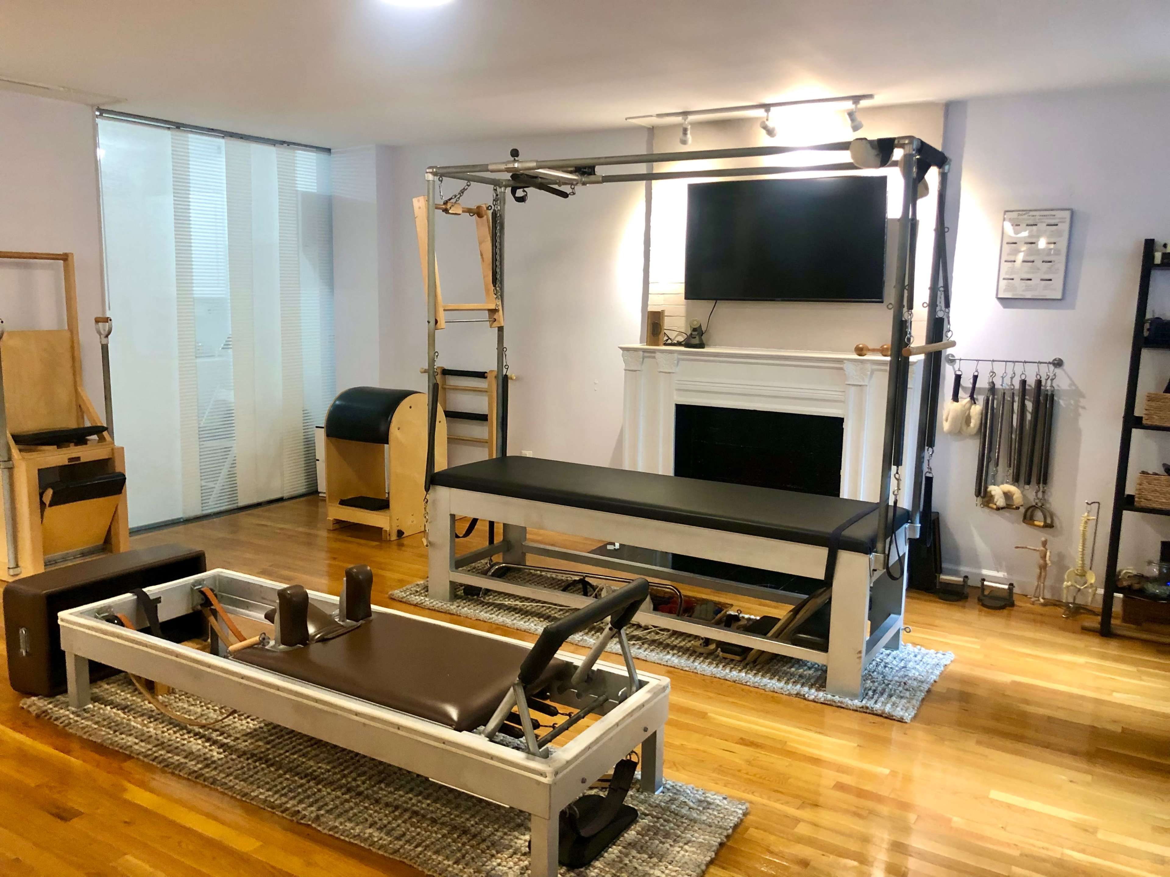 A neatly organized indoor studio featuring a reformer Pilates machine, an elevated Pilates platform, and various fitness equipment along with a television mounted on the wall.