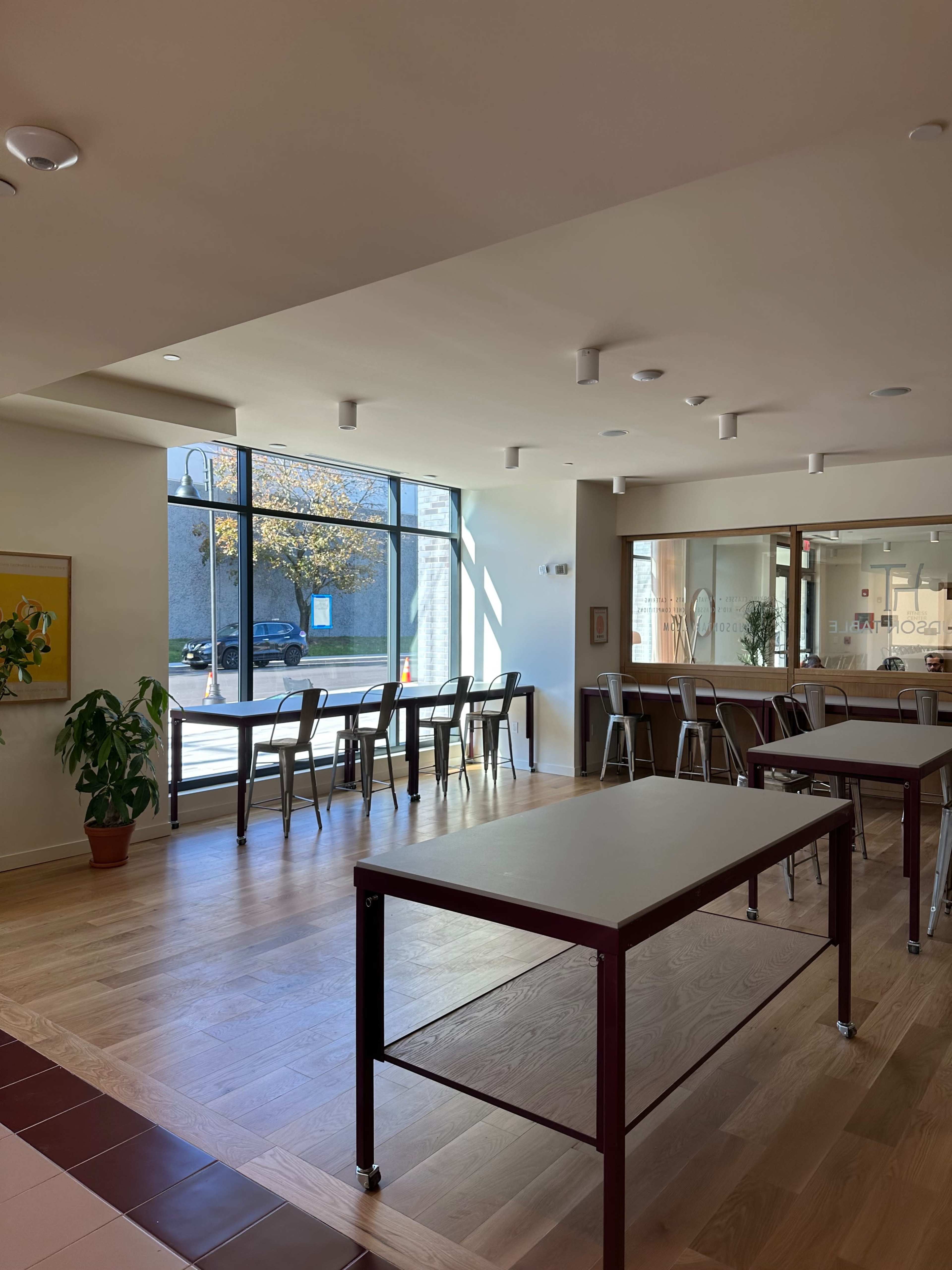 The interior of a modern café features wooden flooring, large windows, and several tables with metal chairs, creating a spacious atmosphere.