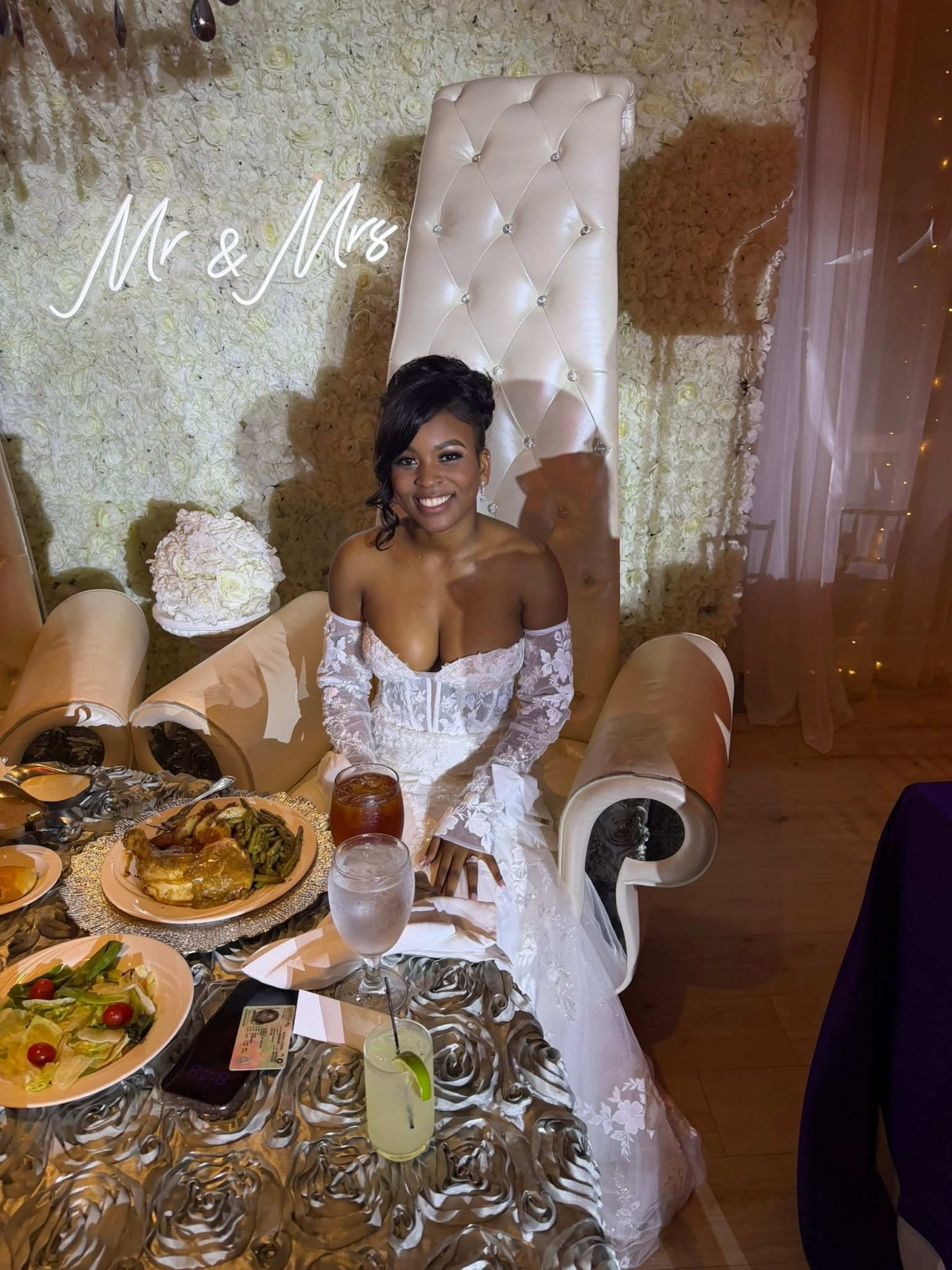 A woman in a white wedding dress sits at a table adorned with food and drinks, in front of a floral backdrop with a decorative chair.