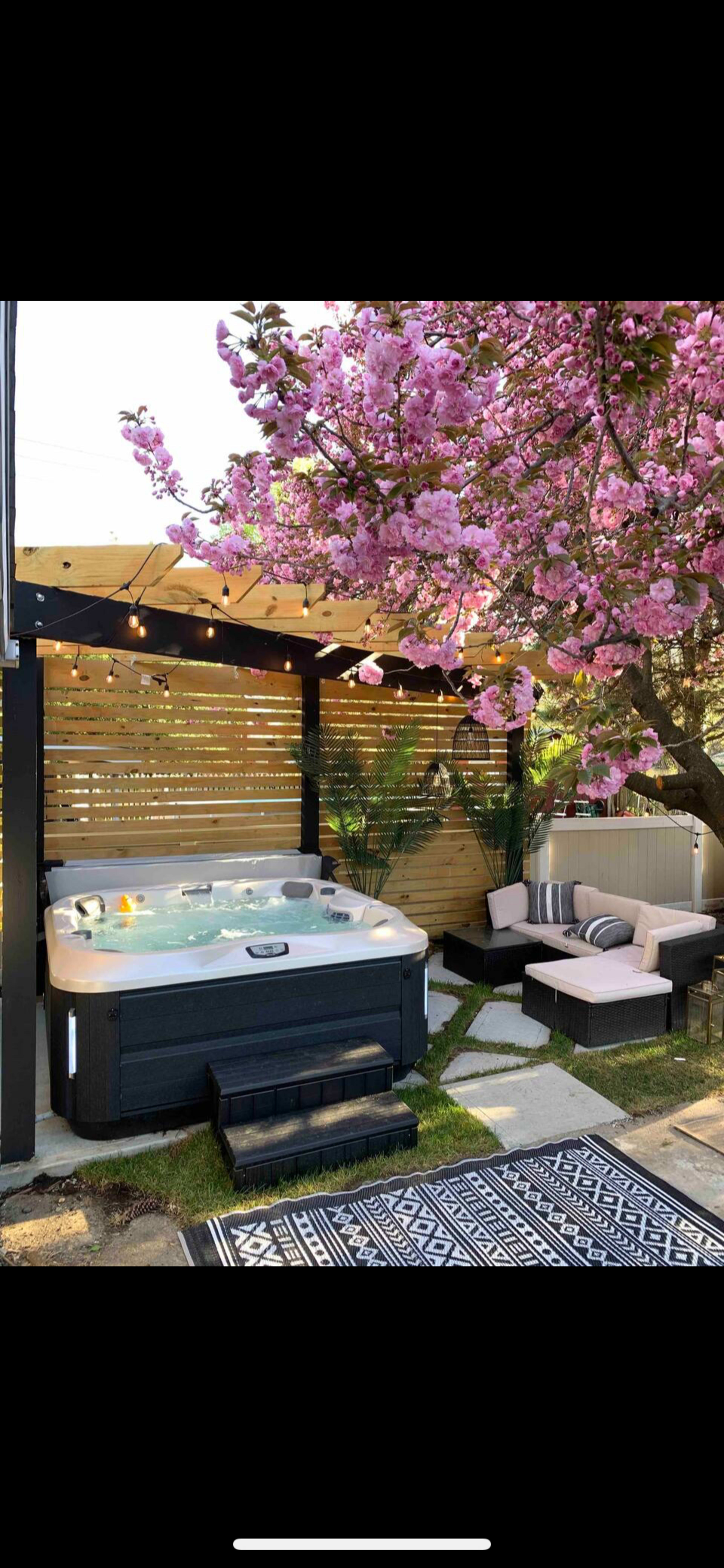 The image shows a backyard space featuring a hot tub under a wooden pergola, surrounded by pink flowering trees and a seating area with cushions.