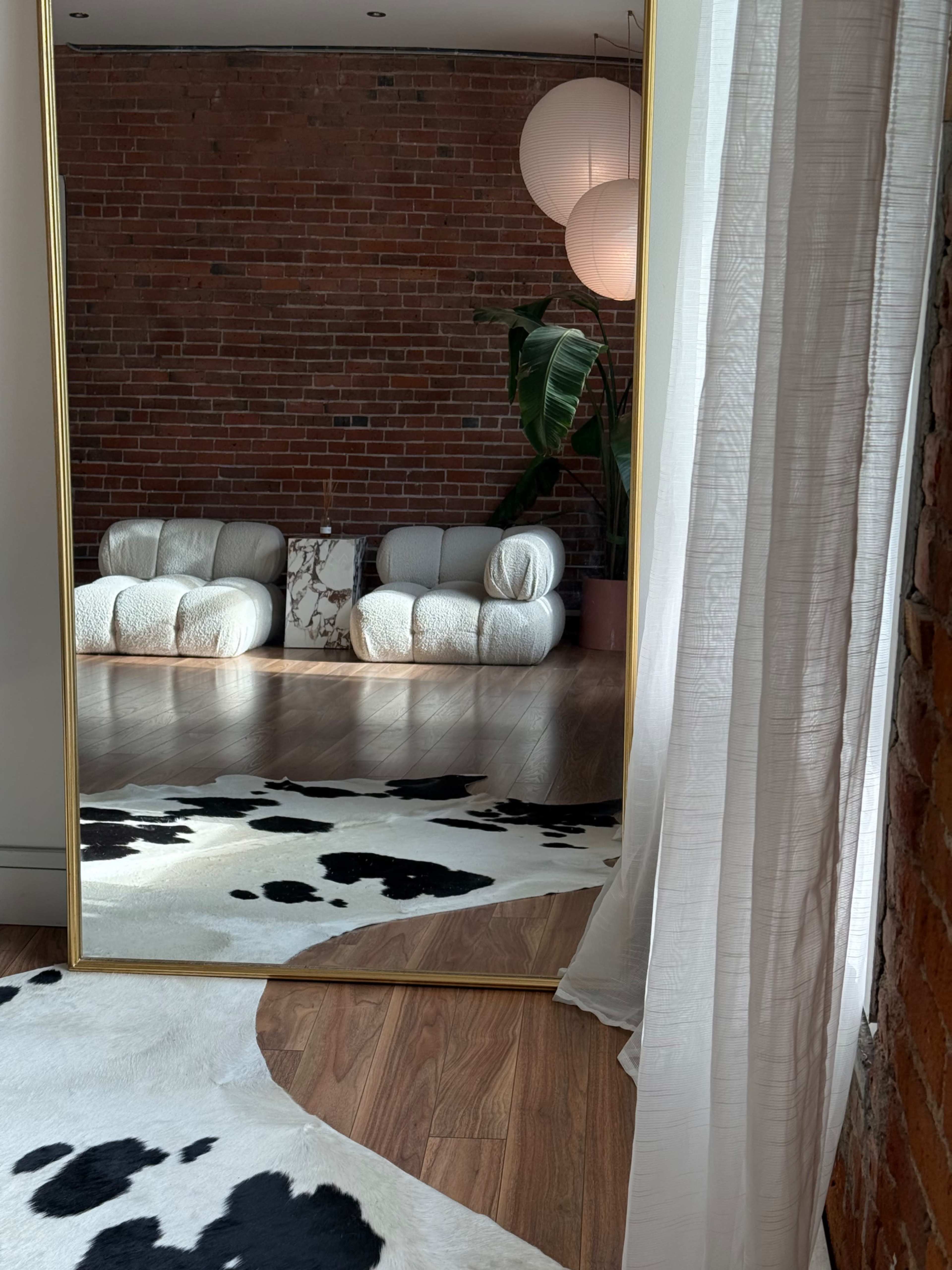 A large floor mirror reflects a cozy seating area with white upholstered chairs and a cowhide rug, set against a brick wall decorated with a potted plant.