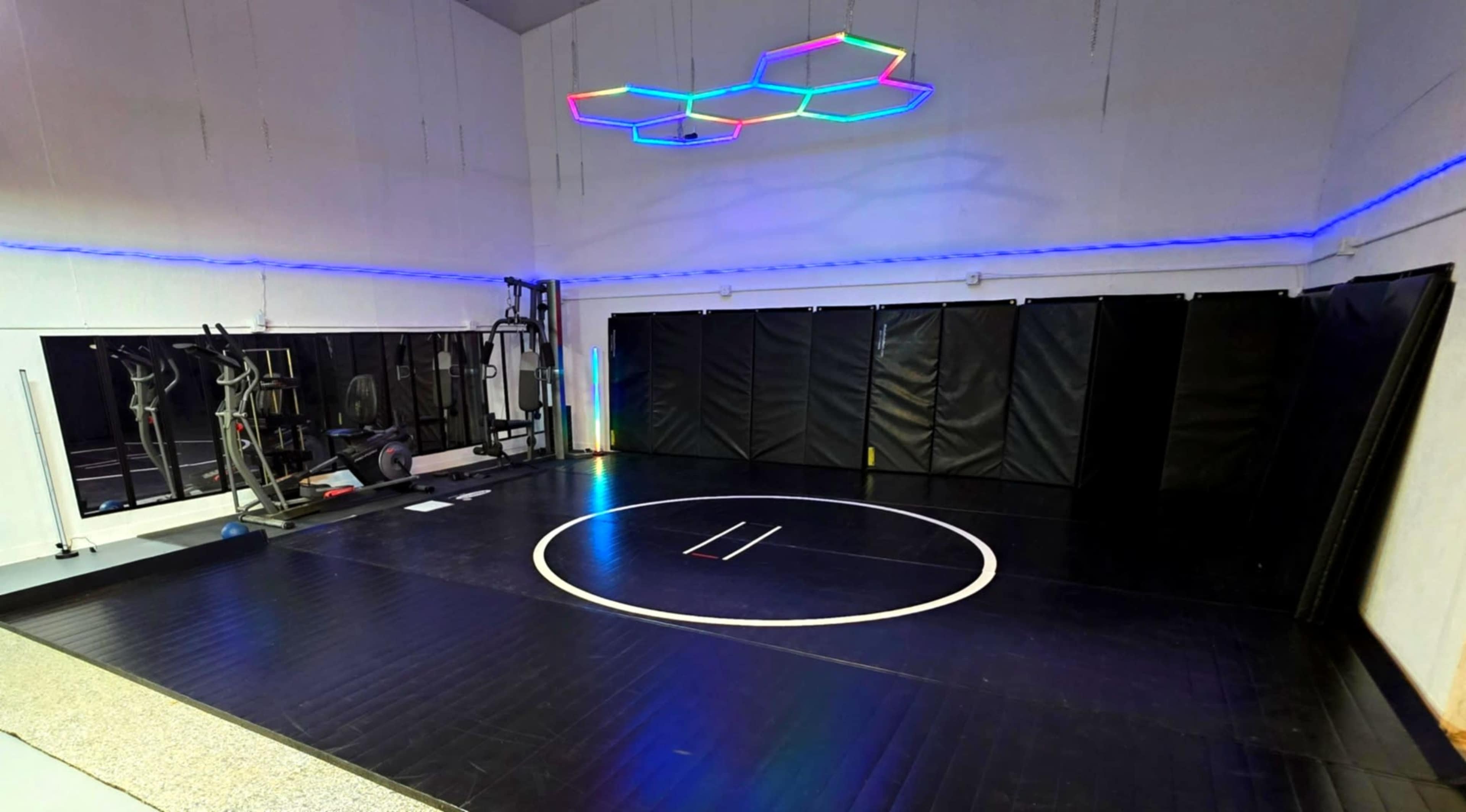 The image shows a spacious wrestling gym featuring black mats, a circular wrestling area marked in white, and colorful LED lights overhead.