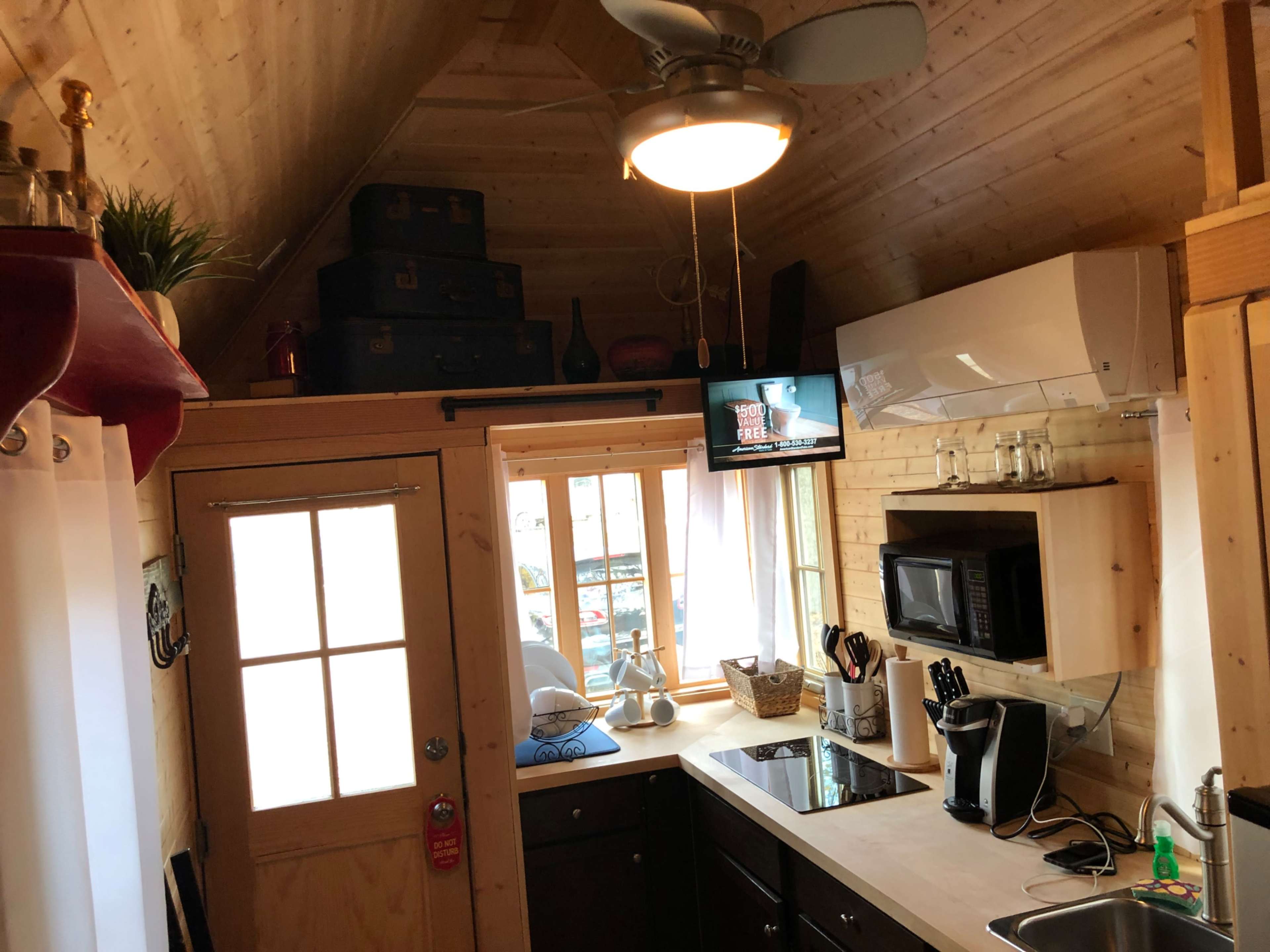 A small kitchen in a wooden cabin featuring a stove, microwave, sink, and a television mounted on the wall.