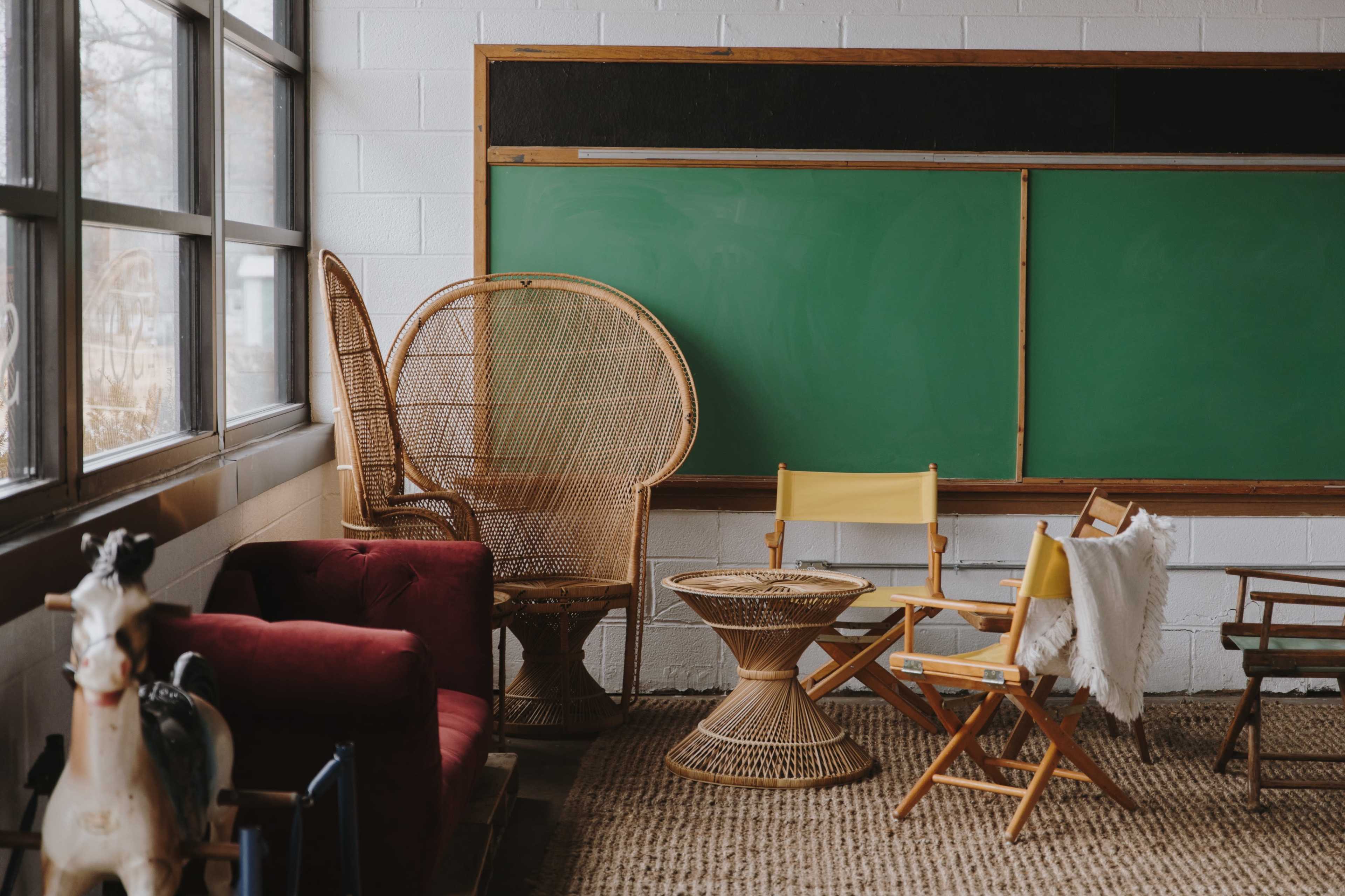 The scene features a classroom with a green chalkboard and various seating options, including wicker chairs and a colorful folding chair, alongside a decorative horse.