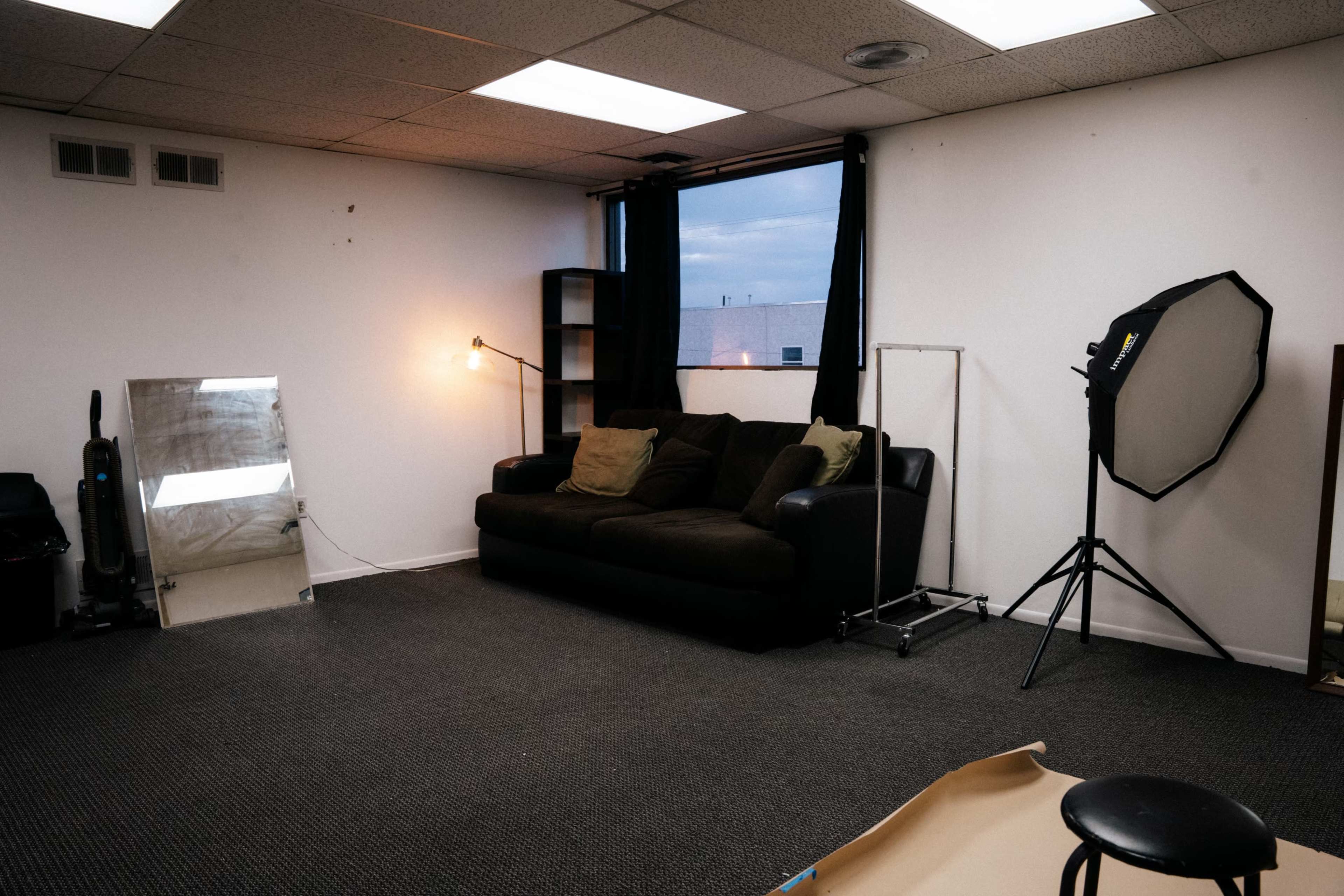 The image shows a sparsely furnished room with a dark sofa, a standing lamp, a mirror, and a photography light setup against a white wall.