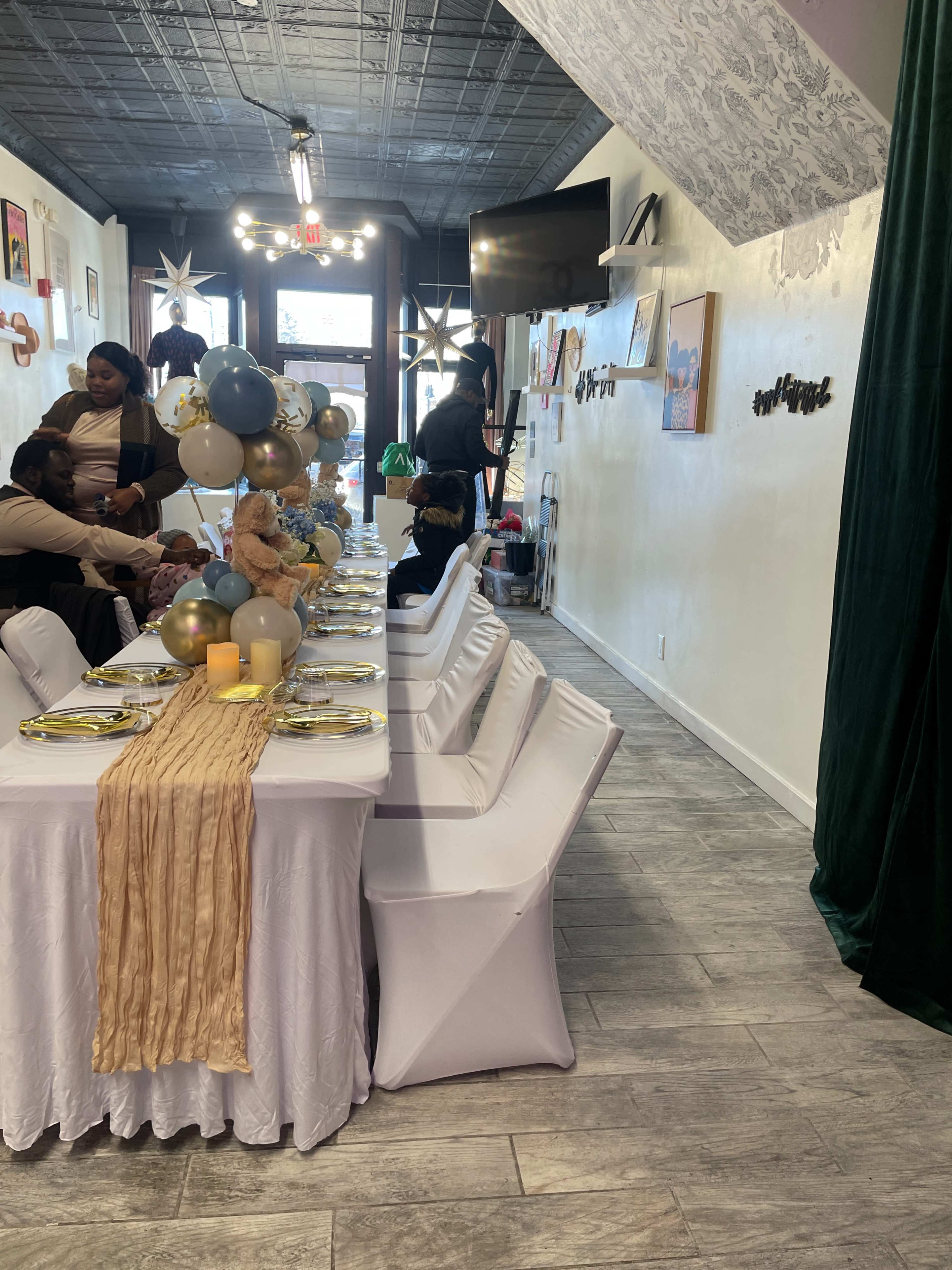 The image shows a long banquet table elegantly set with white tablecloths, golden plates, and decorative balloons in a well-lit event space, with guests seated nearby.
