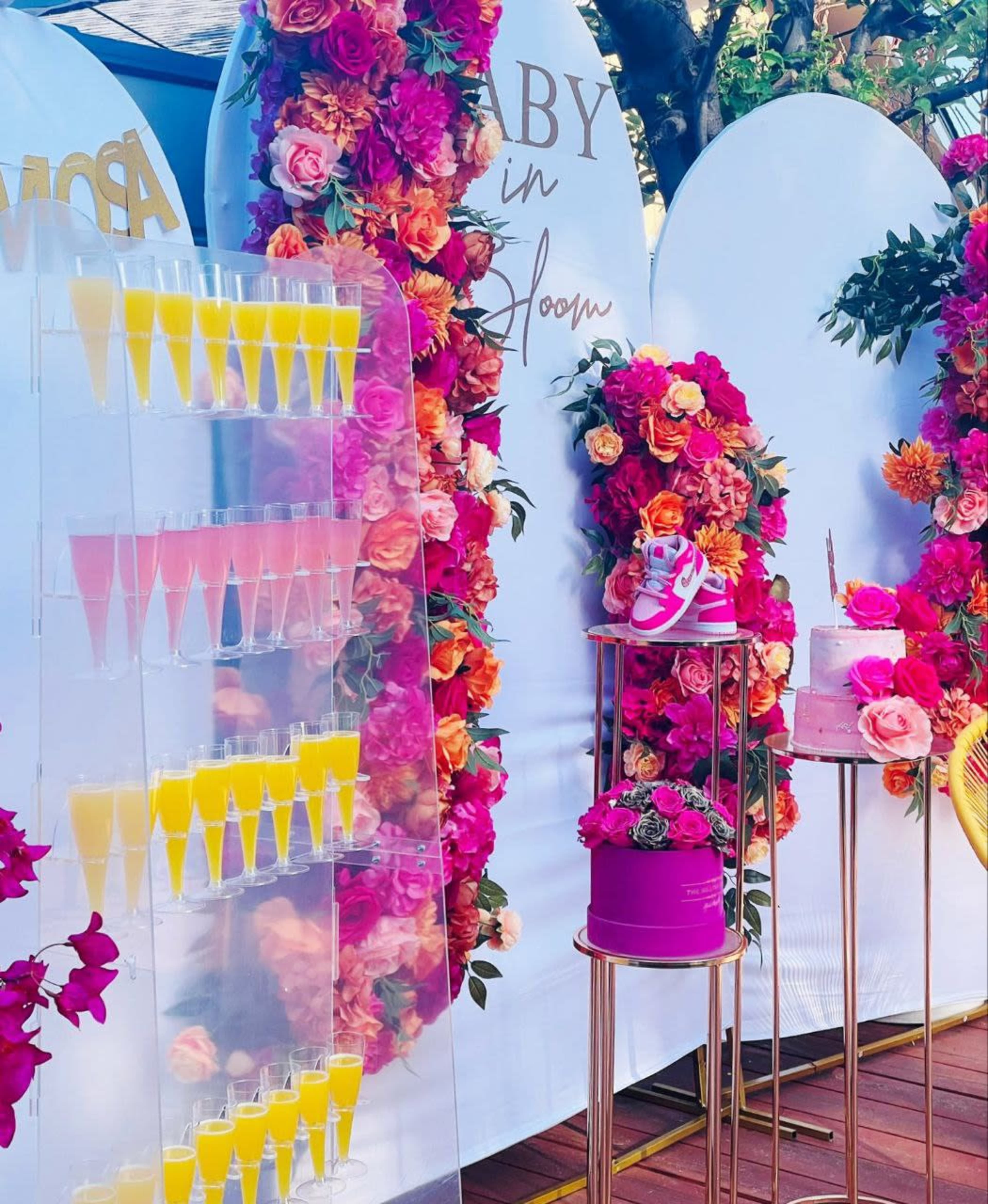 The image features a colorful display for a celebration with a tiered stand of brightly colored drinks, a decorated cake, and floral arrangements in vibrant hues.