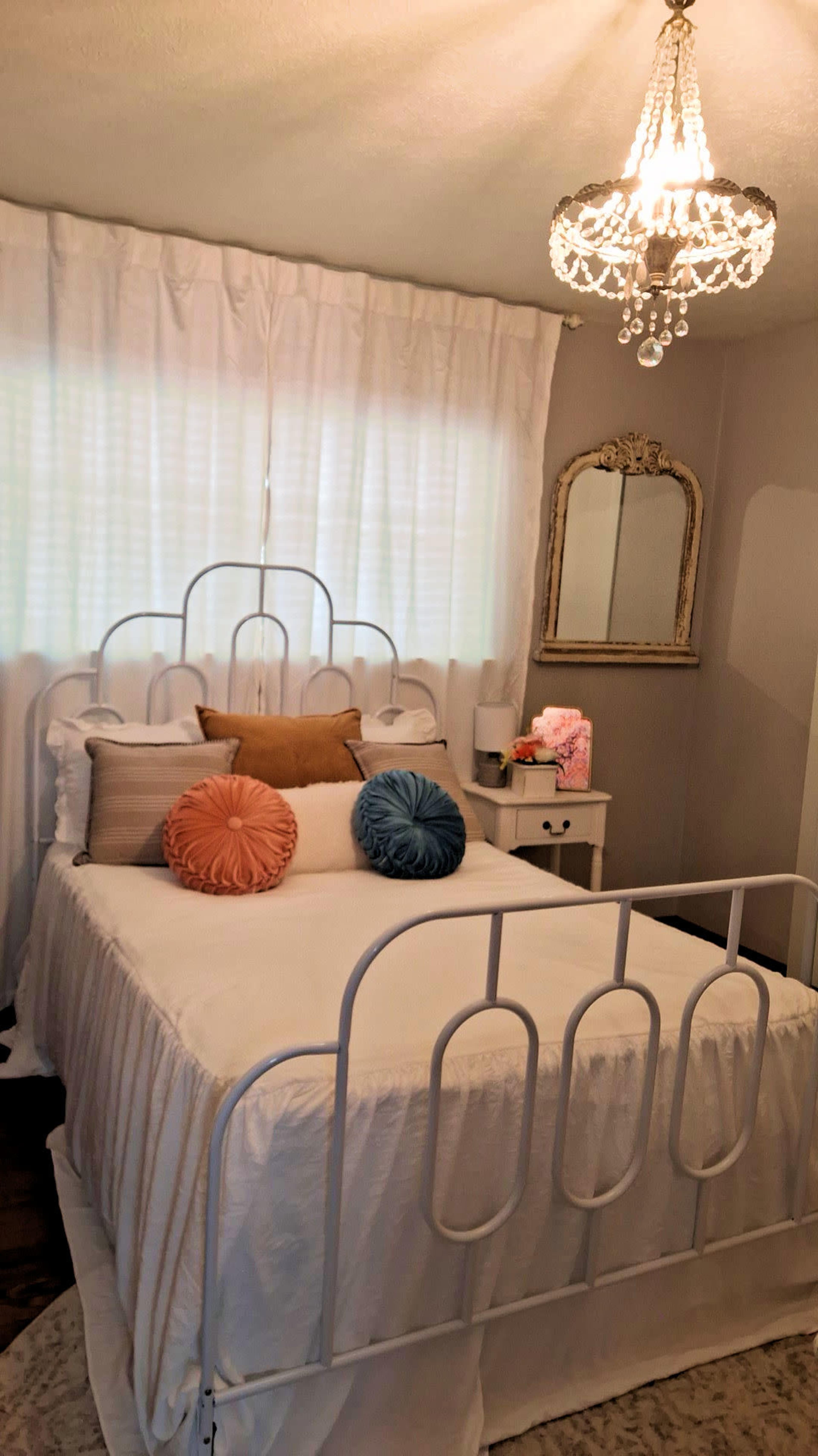 A neatly made bed with decorative pillows in a well-lit bedroom featuring a chandelier and a mirror on the wall.