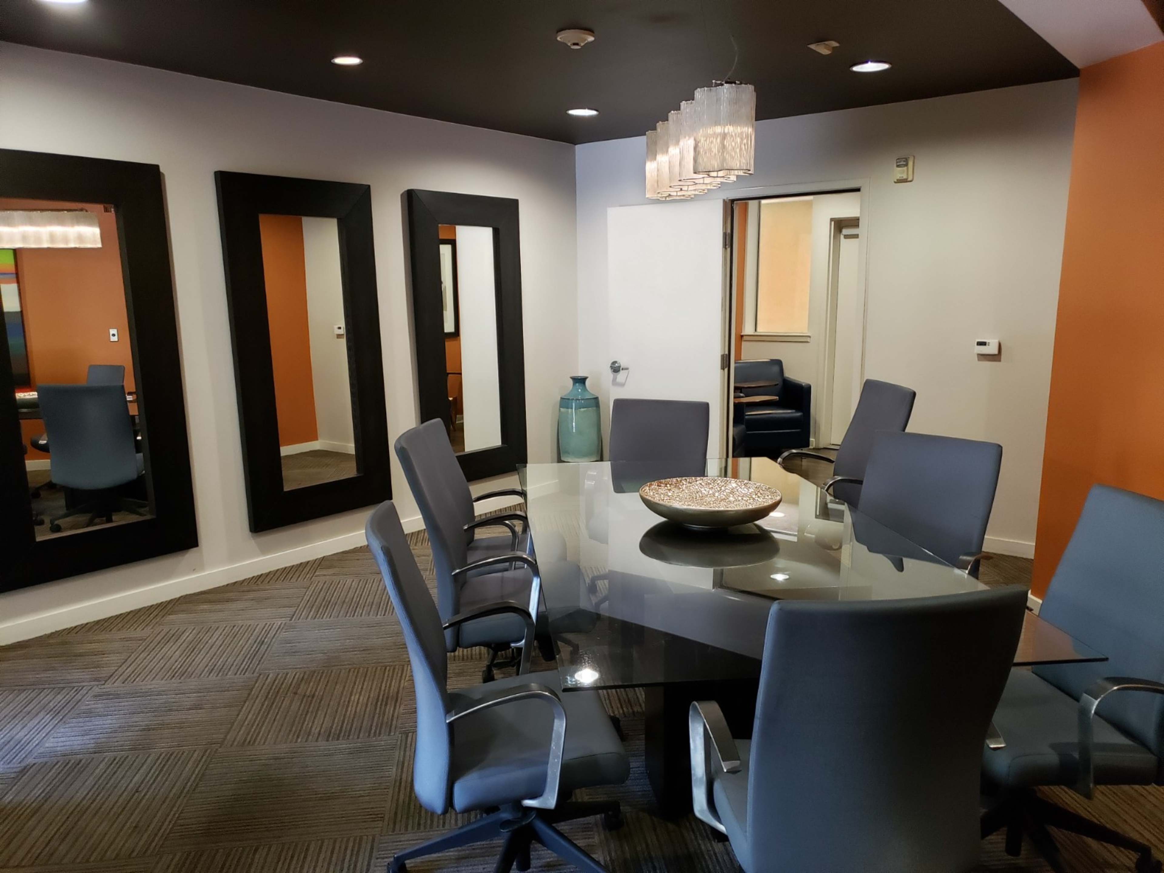 A conference room features a glass table surrounded by six gray chairs, with large mirrors on the walls and an orange accent wall.