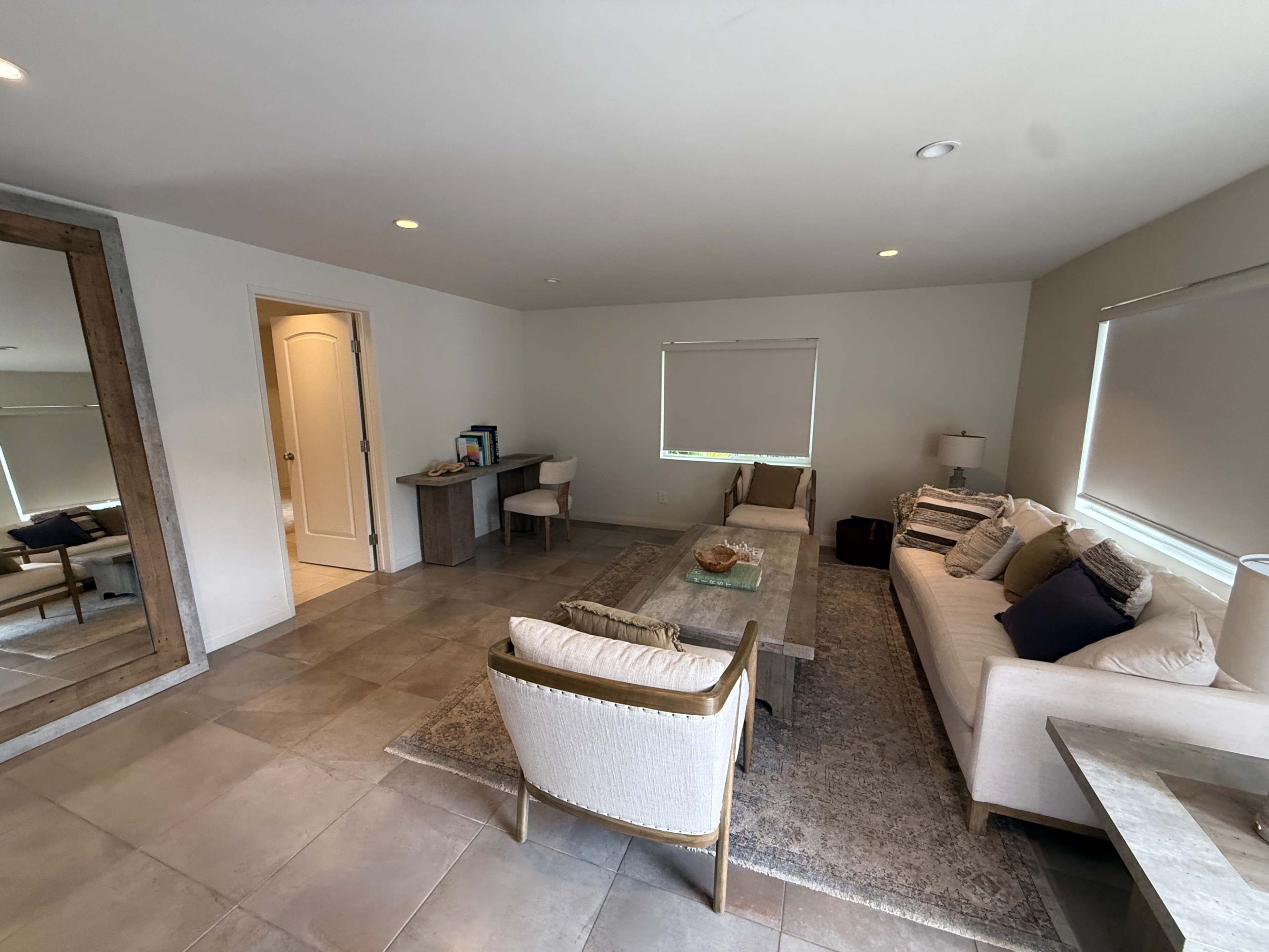 The image shows a spacious living room with a sofa, chairs, coffee table, and a large mirror, featuring neutral-colored walls and tiled flooring.