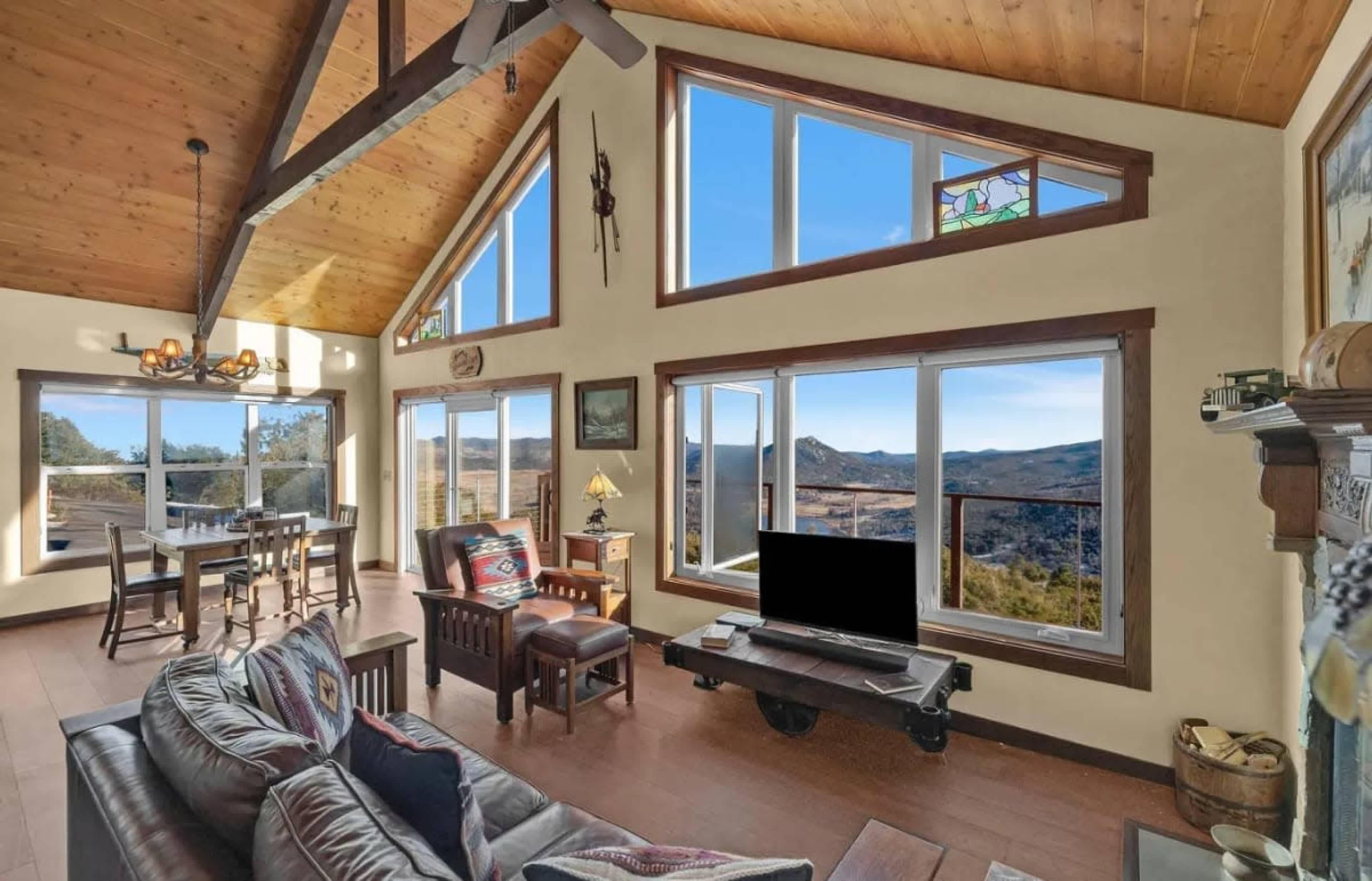 The image shows a modern living room with large windows offering a view of a mountainous landscape, wooden ceilings, and contemporary furniture.