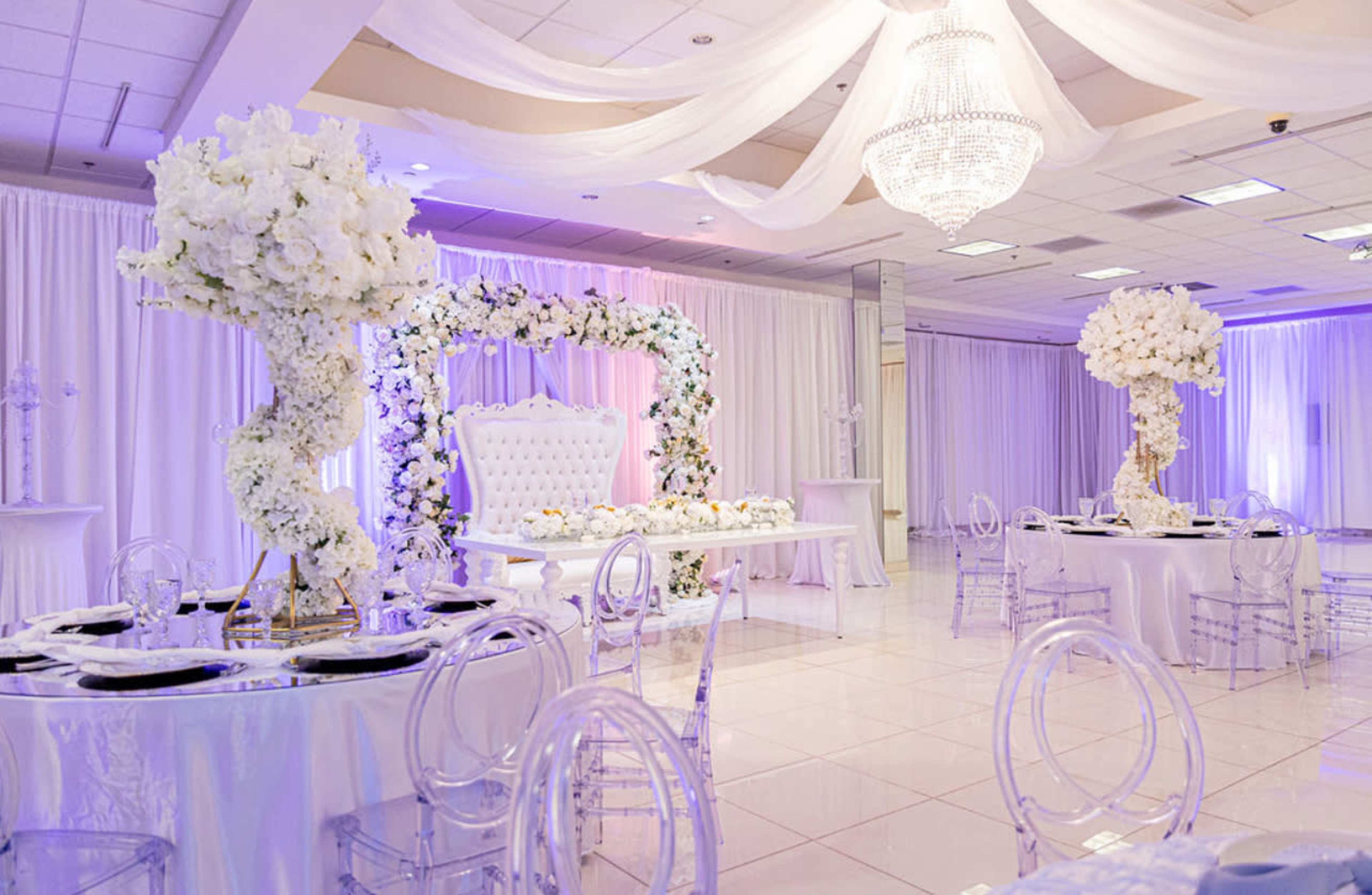 The image shows a beautifully decorated event space with white drapery, elegant floral arrangements, and crystal chandeliers.