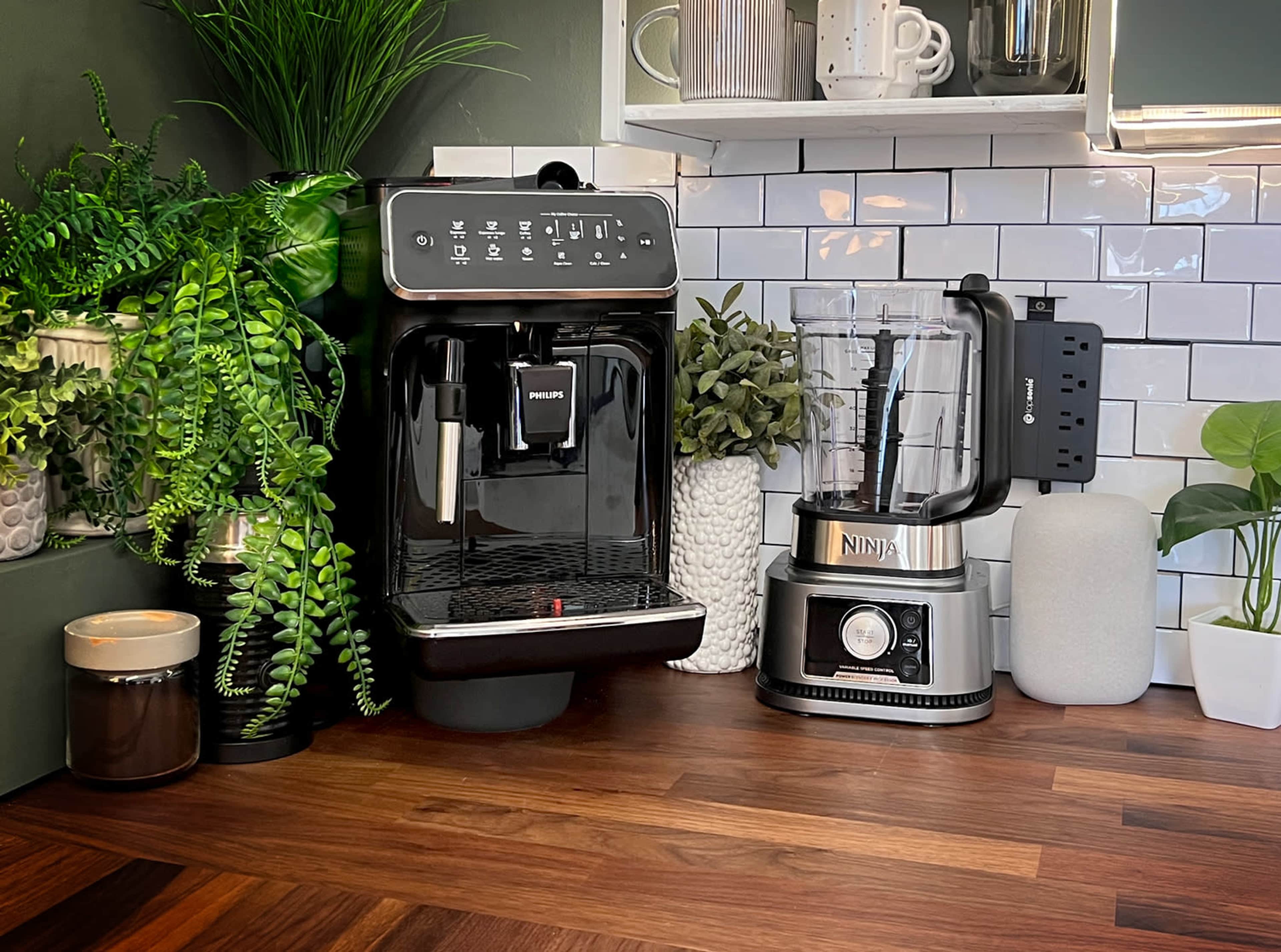 A modern kitchen countertop displays a black Philips coffee maker next to a silver Ninja blender, surrounded by various plants and kitchenware.