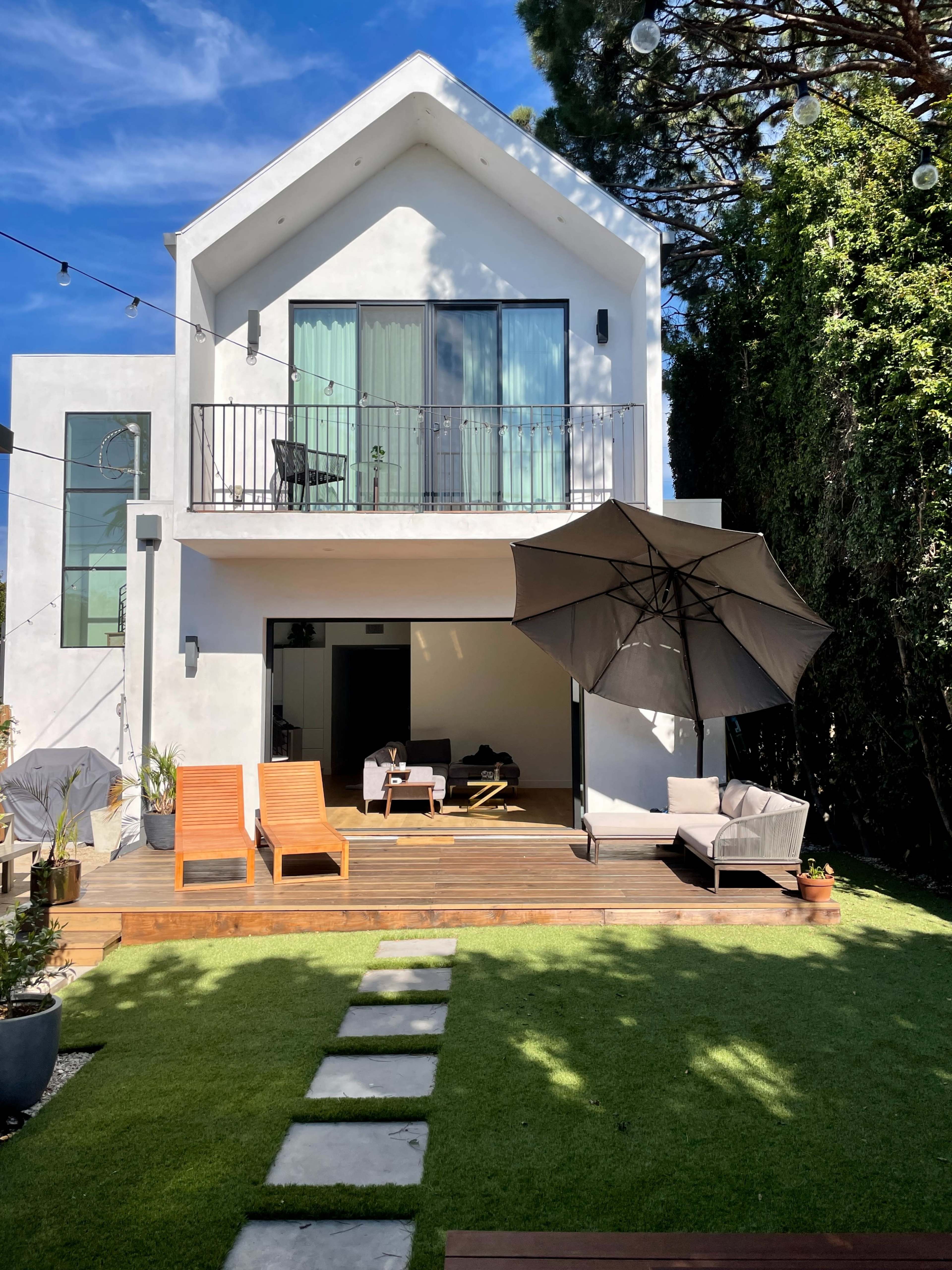 The image shows a modern two-story house with a balcony, surrounded by a green lawn and featuring wooden loungers and an umbrella on a patio.
