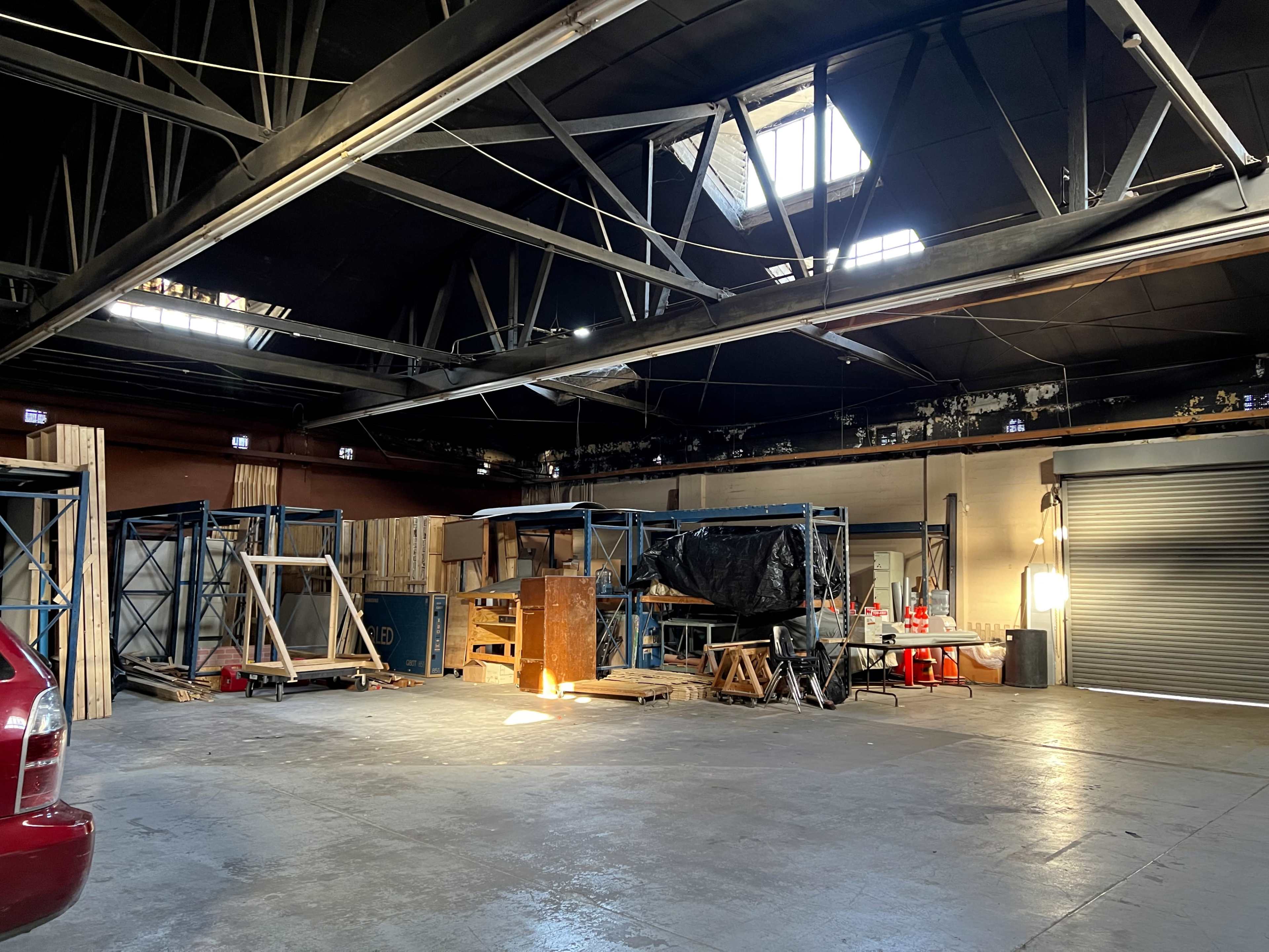 A large industrial space with storage shelves, wooden materials, and a red car parked nearby under natural light from a window.