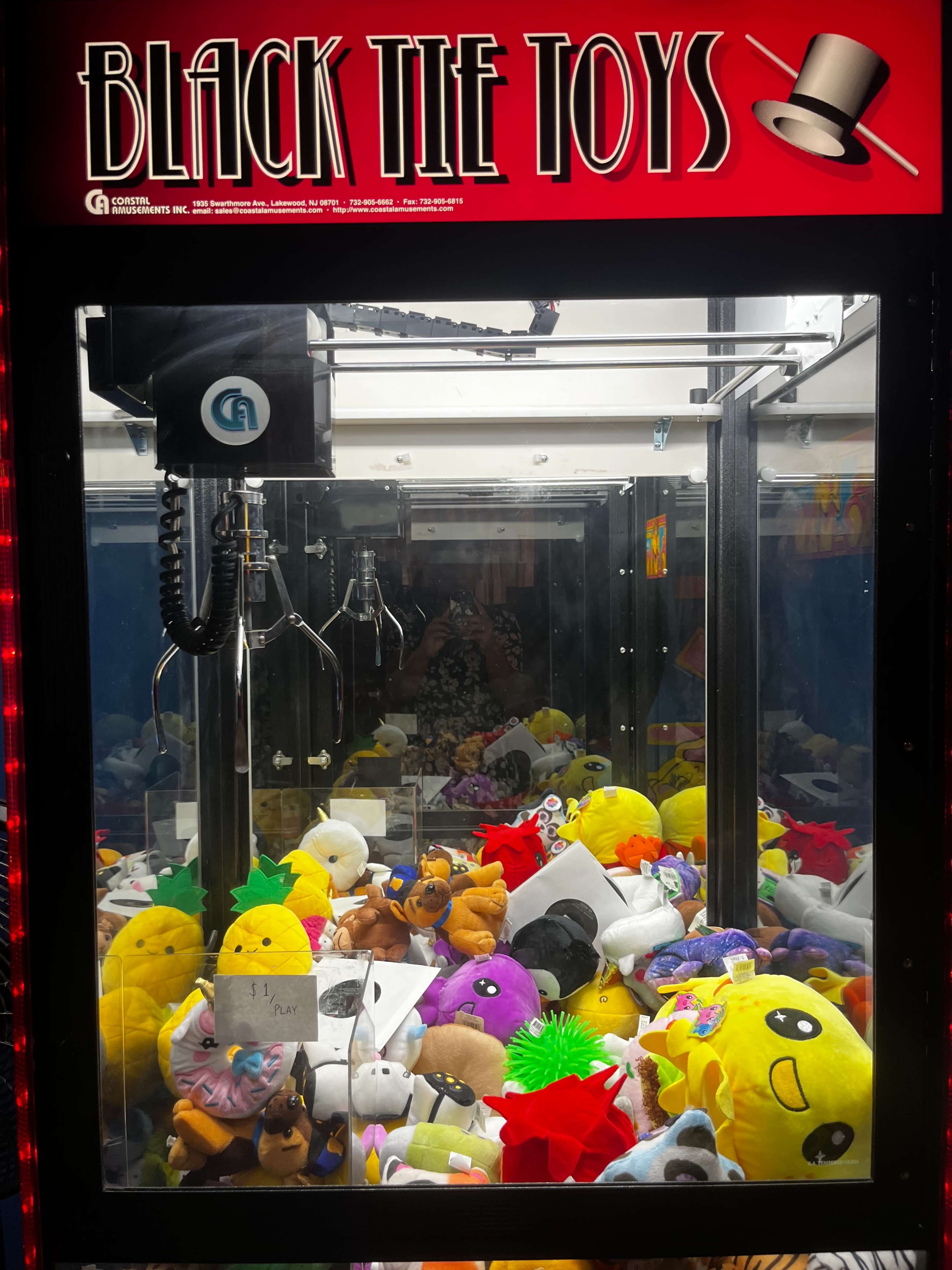 A claw machine filled with various plush toys, including animals and colorful characters, under a sign that reads "BLACK TIE TOYS."