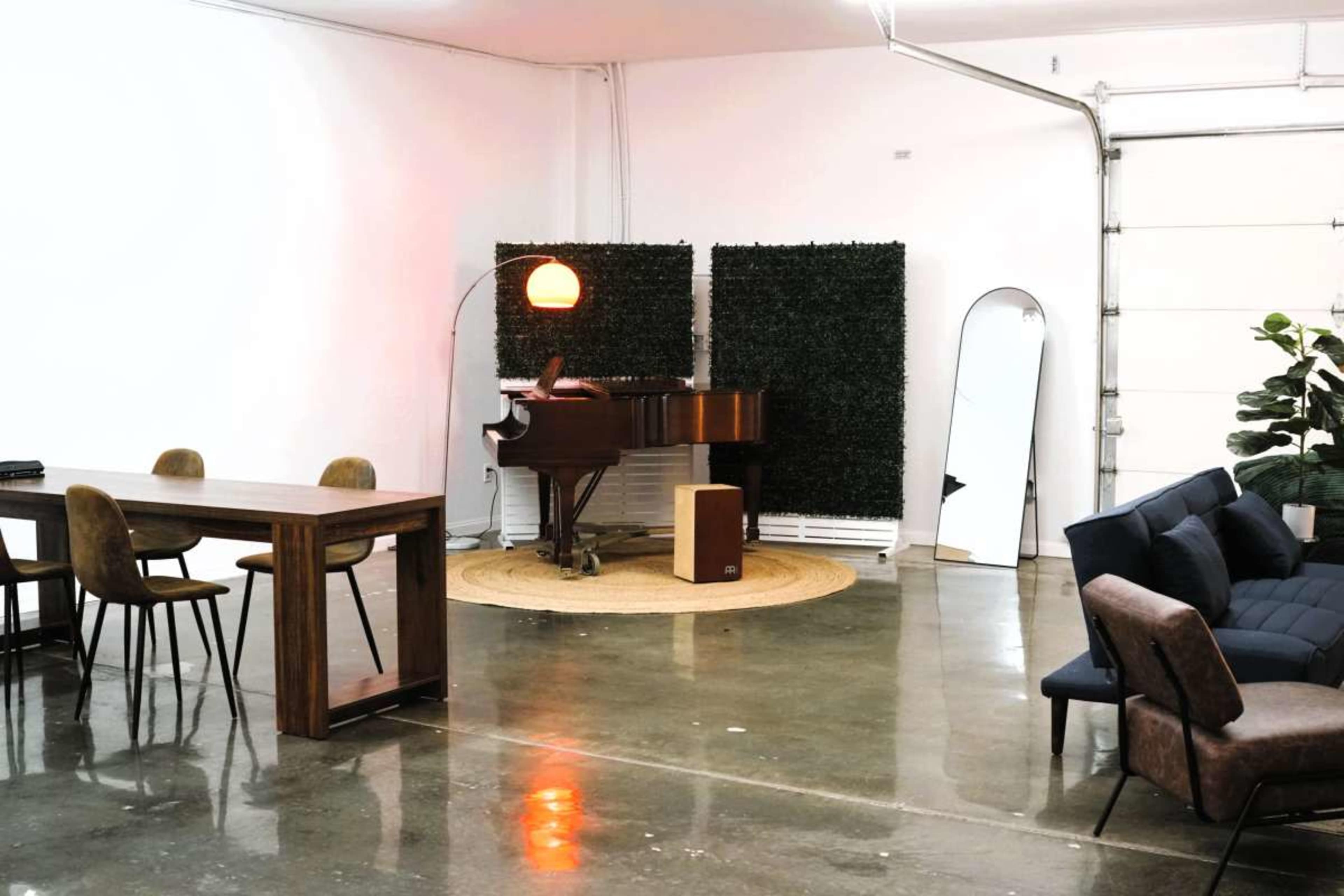 The image shows a modern indoor space featuring a piano, a dining table with chairs, a large mirror, and a sofa, all set against a backdrop of greenery and a garage door.