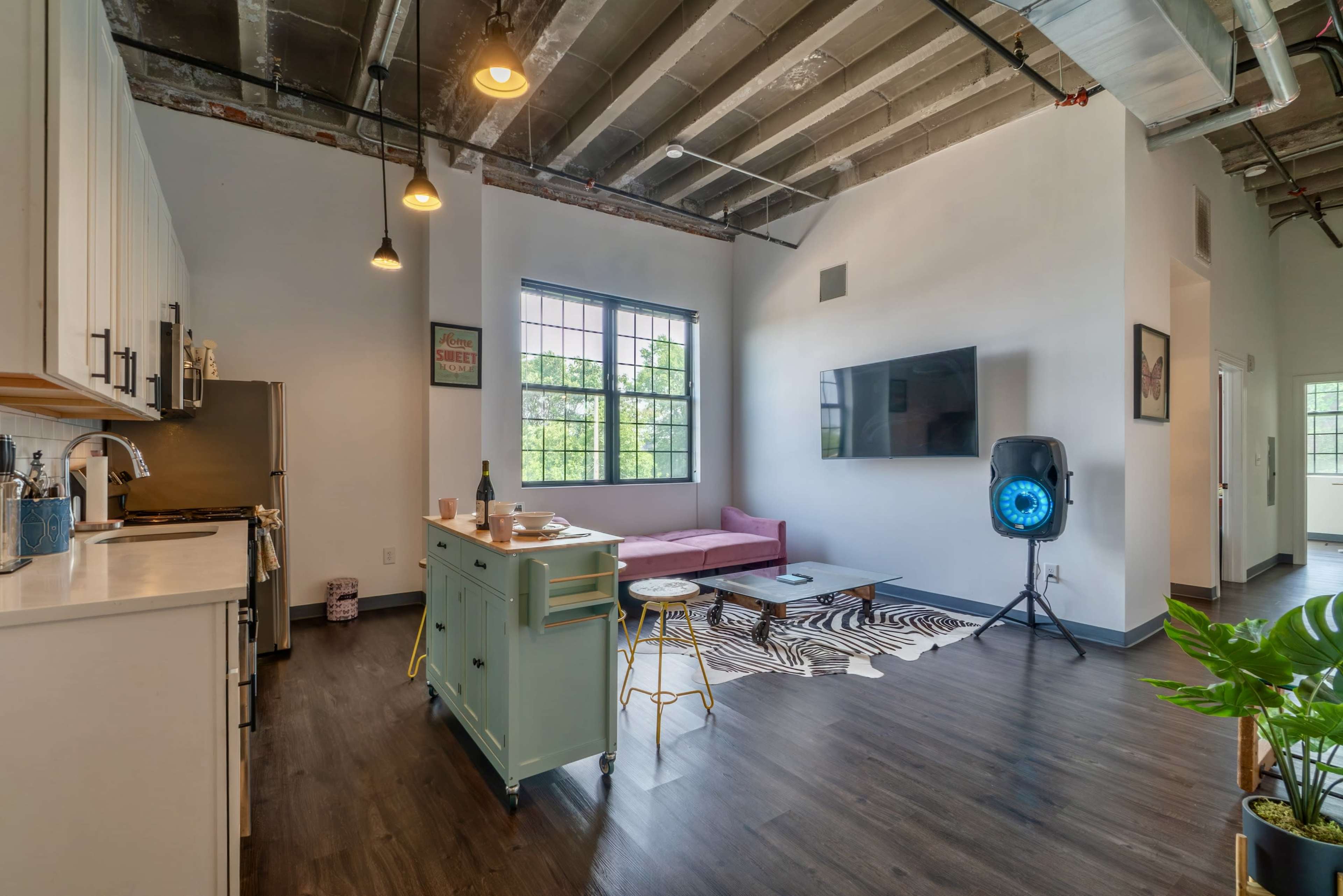 A modern living space features an open kitchen with a dining area, a pink sofa, a television mounted on the wall, and a decorative rug on the floor.
