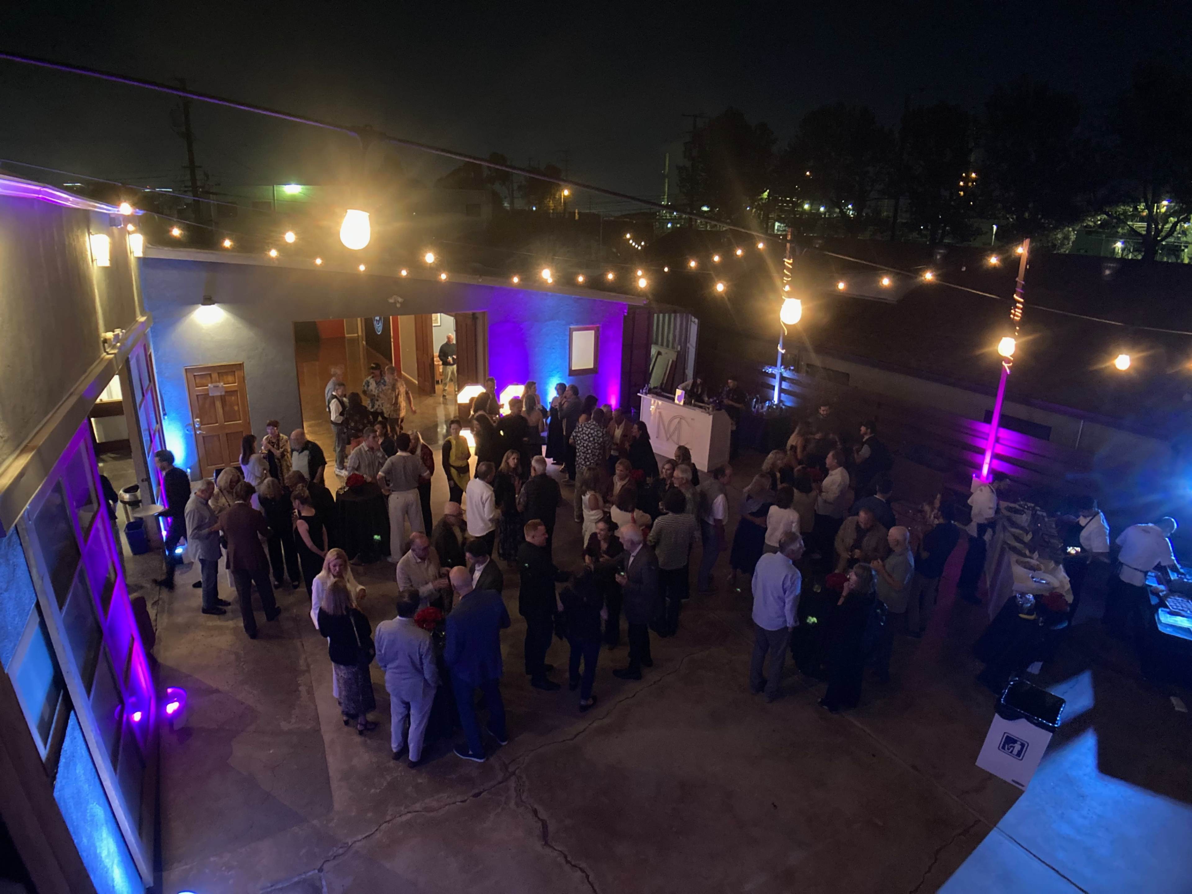 A large group of people mingles under string lights in an outdoor space at night.