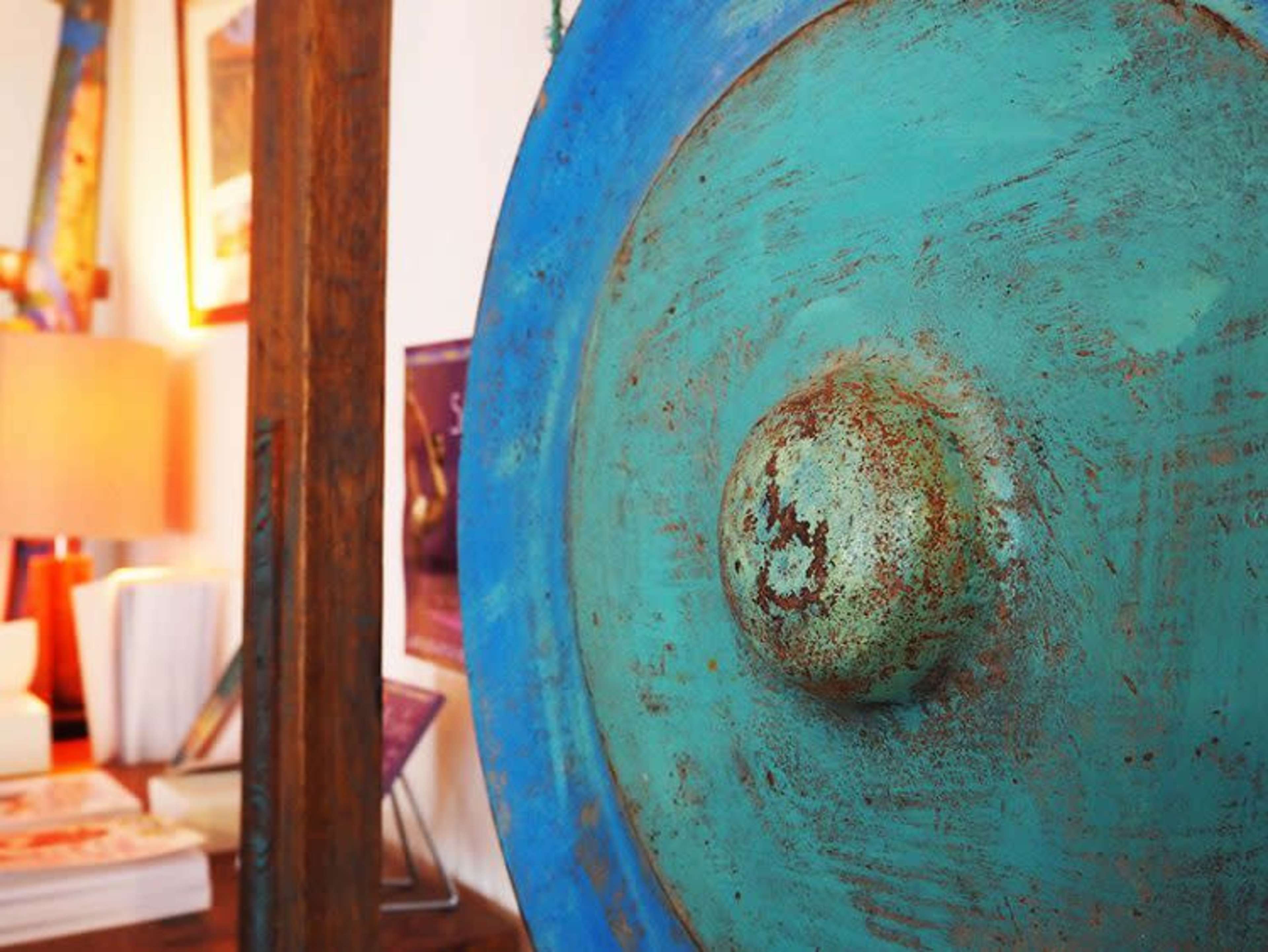 A close-up of a weathered turquoise gong with a rusted surface, positioned near a wooden frame and a blurred background of a cozy interior space.