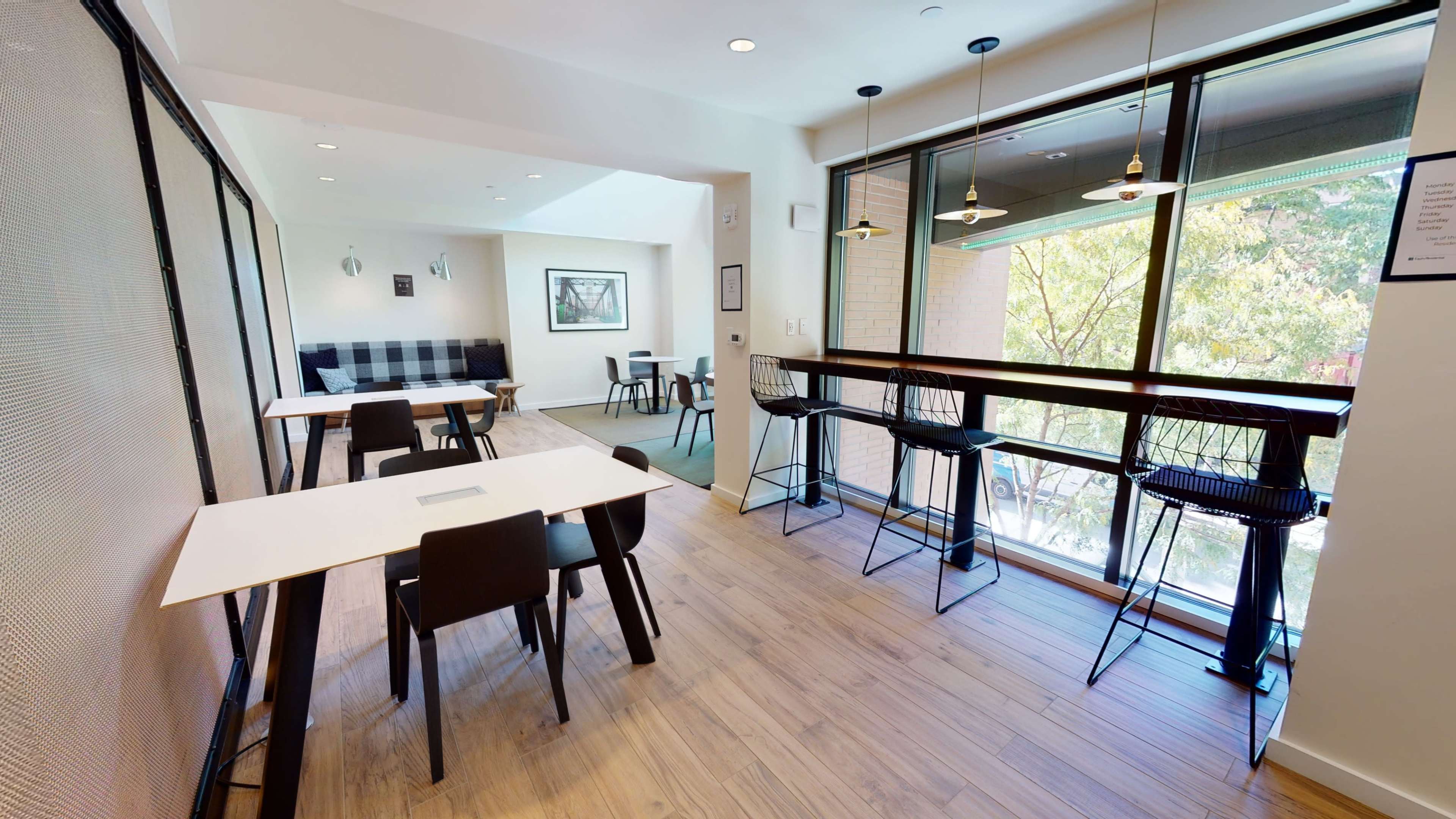 A modern, well-lit workspace features tables with chairs, a seating area with a sofa, and large windows overlooking greenery.