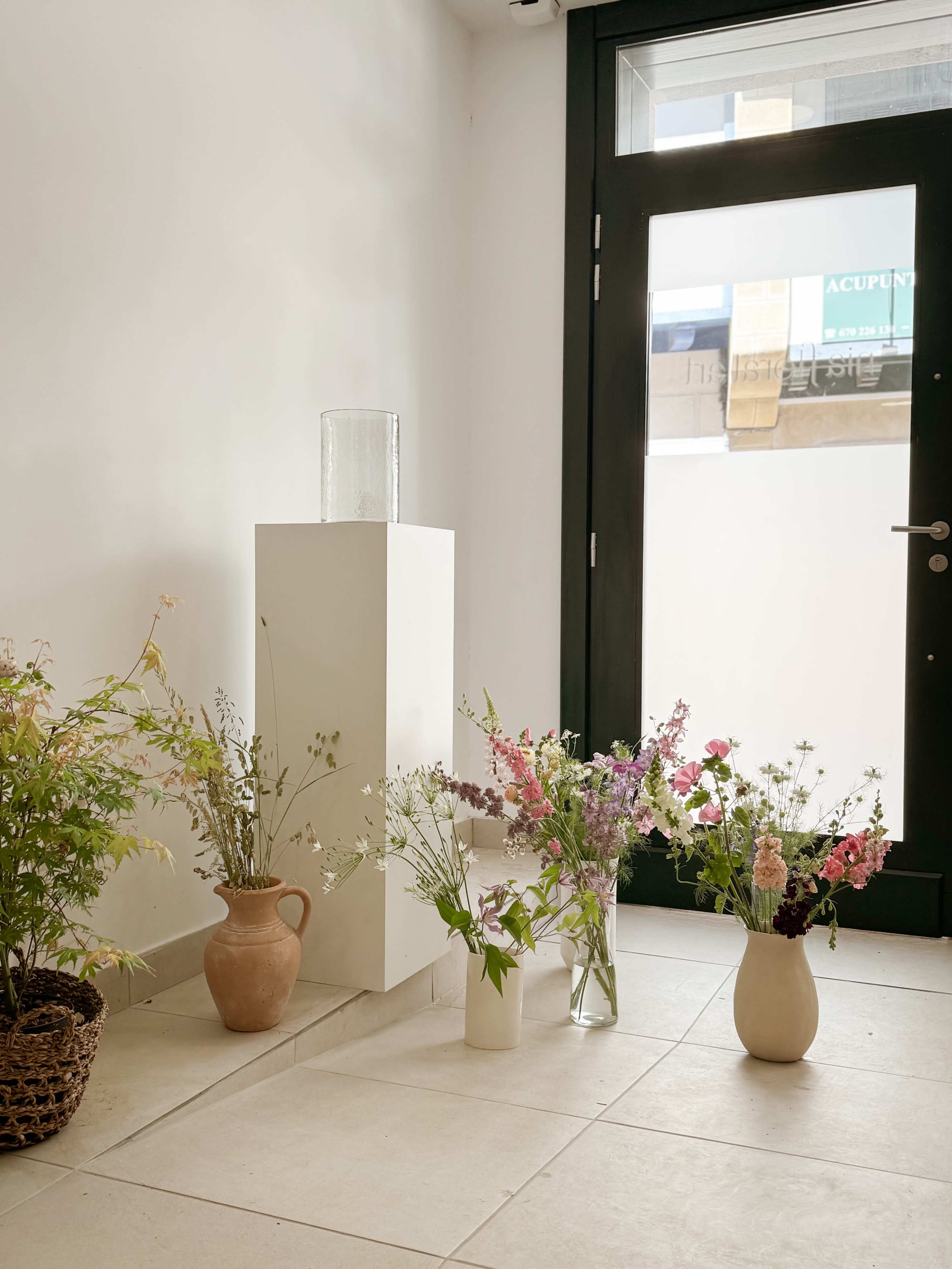 The image shows a variety of potted flowers and plants arranged next to a pedestal in a room with a large glass door.