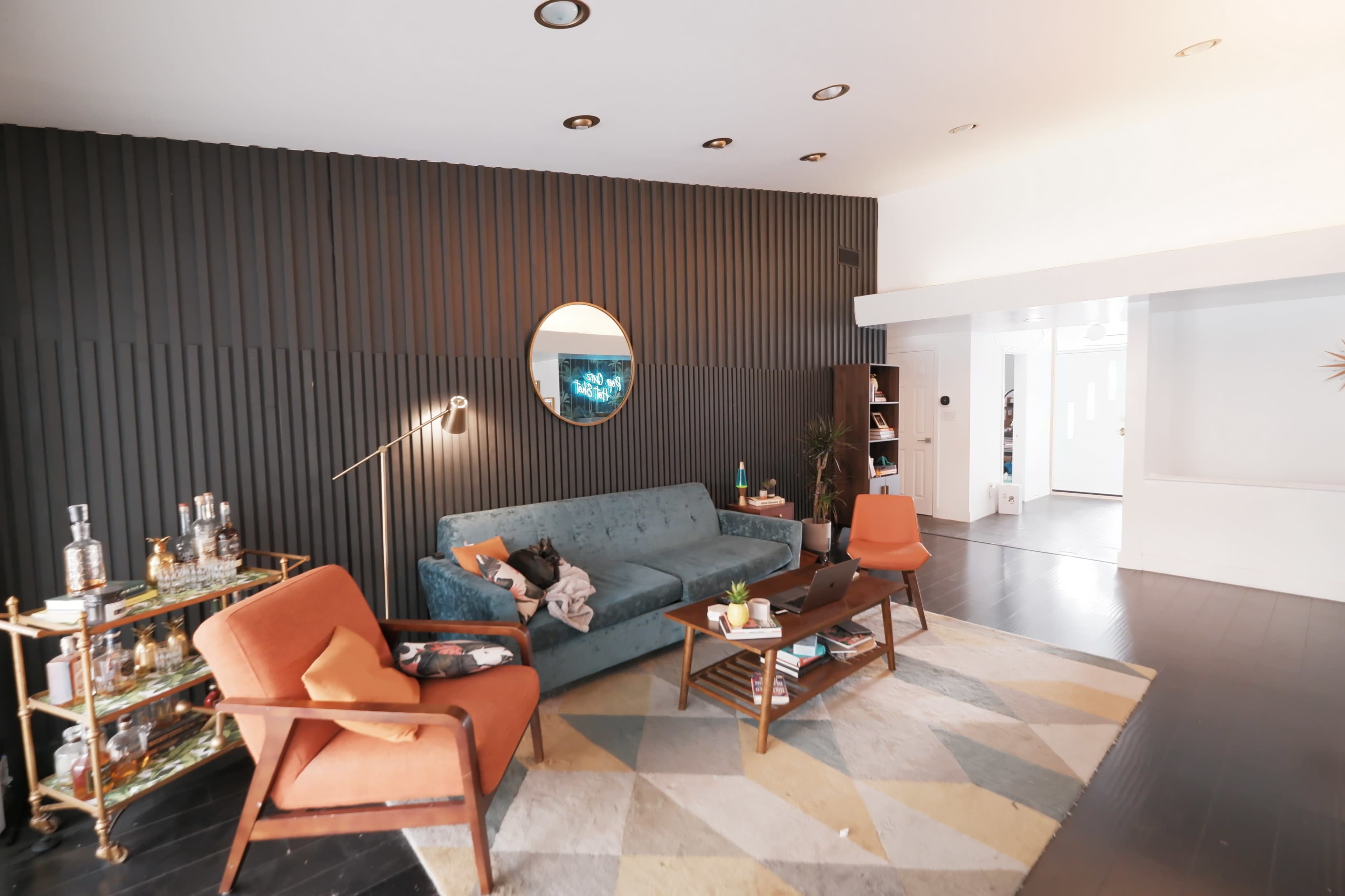 A modern living room featuring a blue sofa, two orange chairs, a circular mirror on the wall, and a patterned area rug.