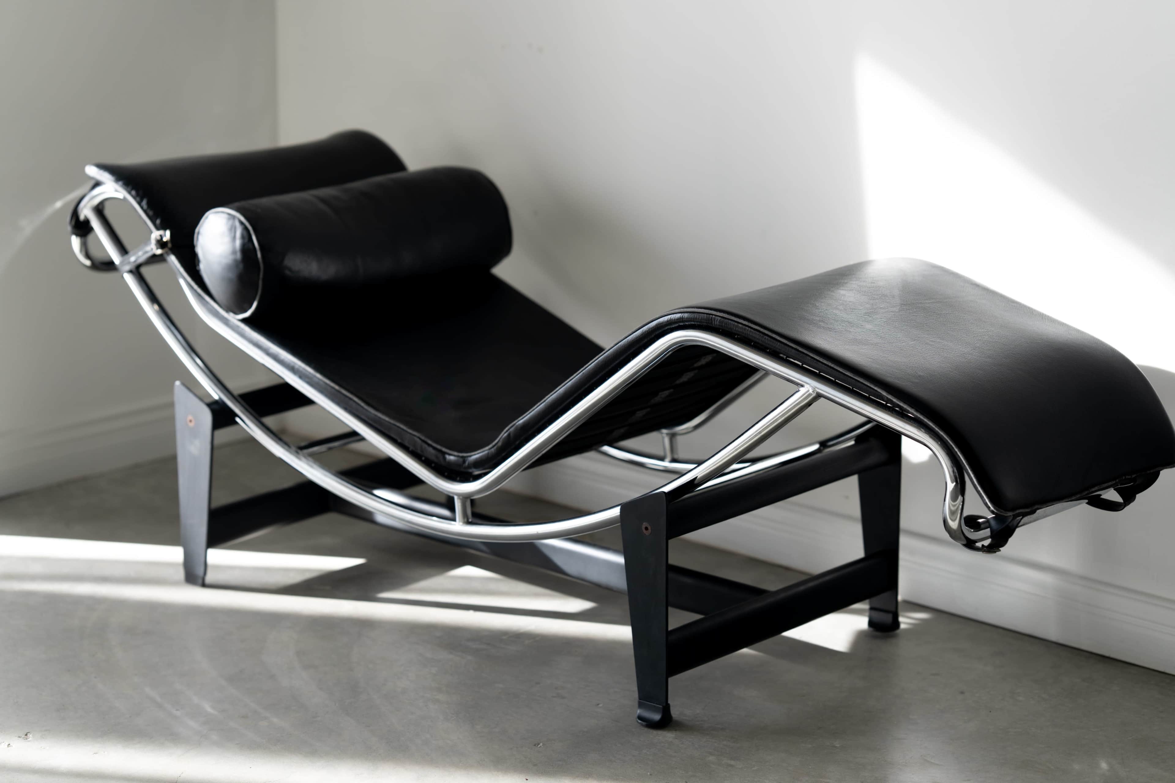 The image shows a modern black leather chaise lounge with a sleek chrome frame positioned against a light wall.