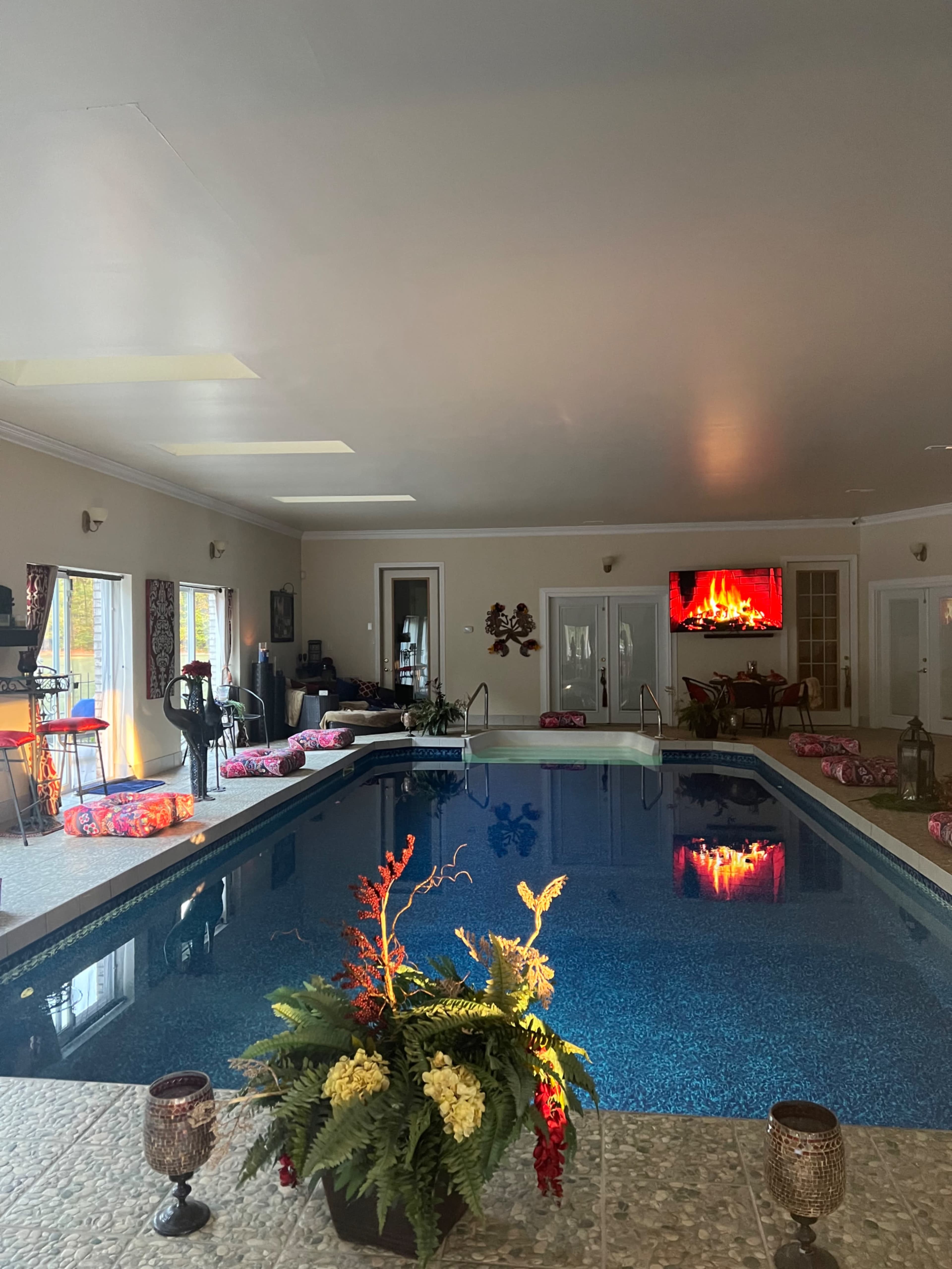 An indoor pool area with a large swimming pool, colorful seating arrangements, and a television displaying a fireplace.