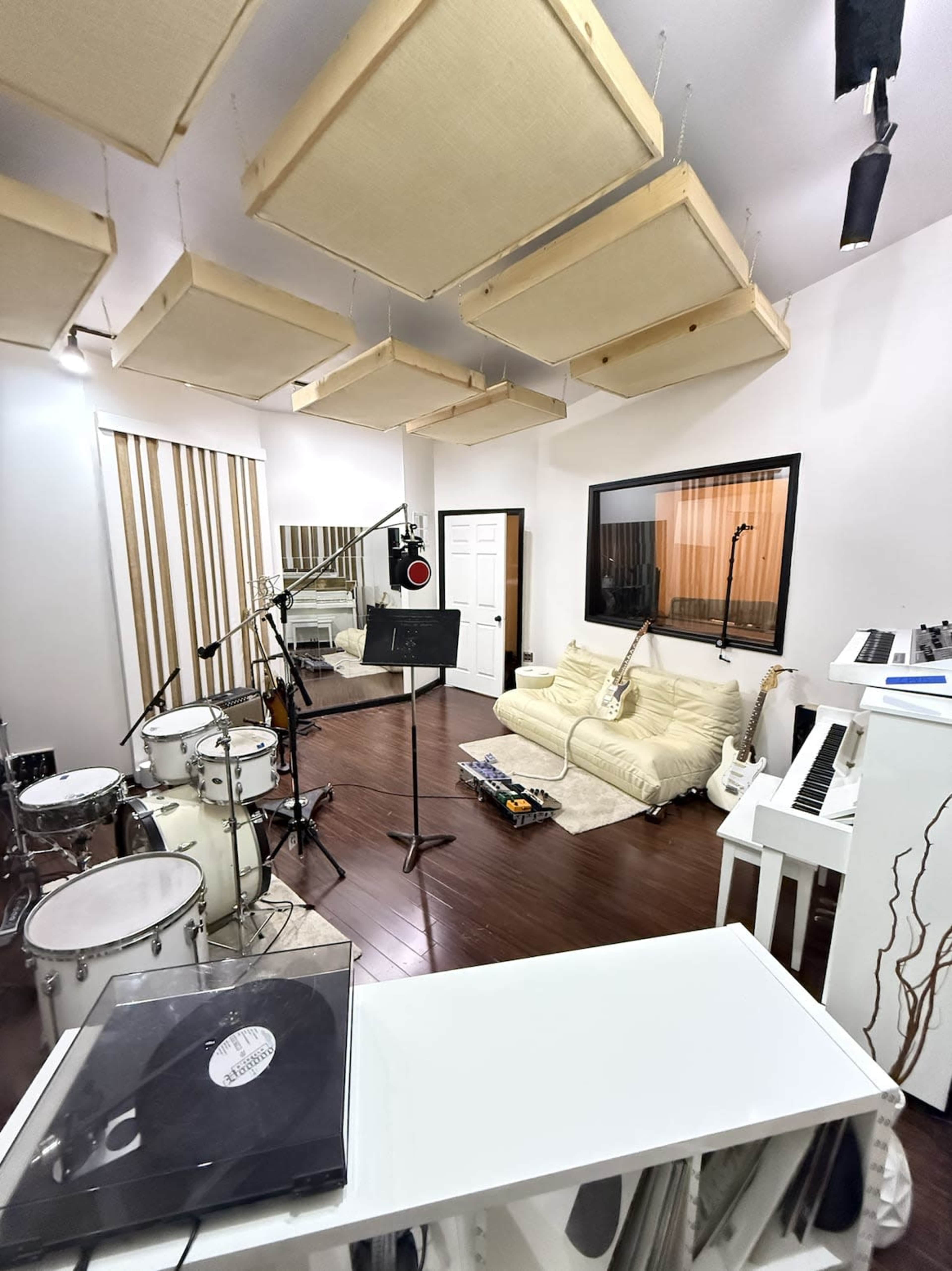 The image shows a modern music studio featuring a drum set, a white piano, a microphone stand, and various instruments arranged in a spacious room with soundproofing panels and wood flooring.
