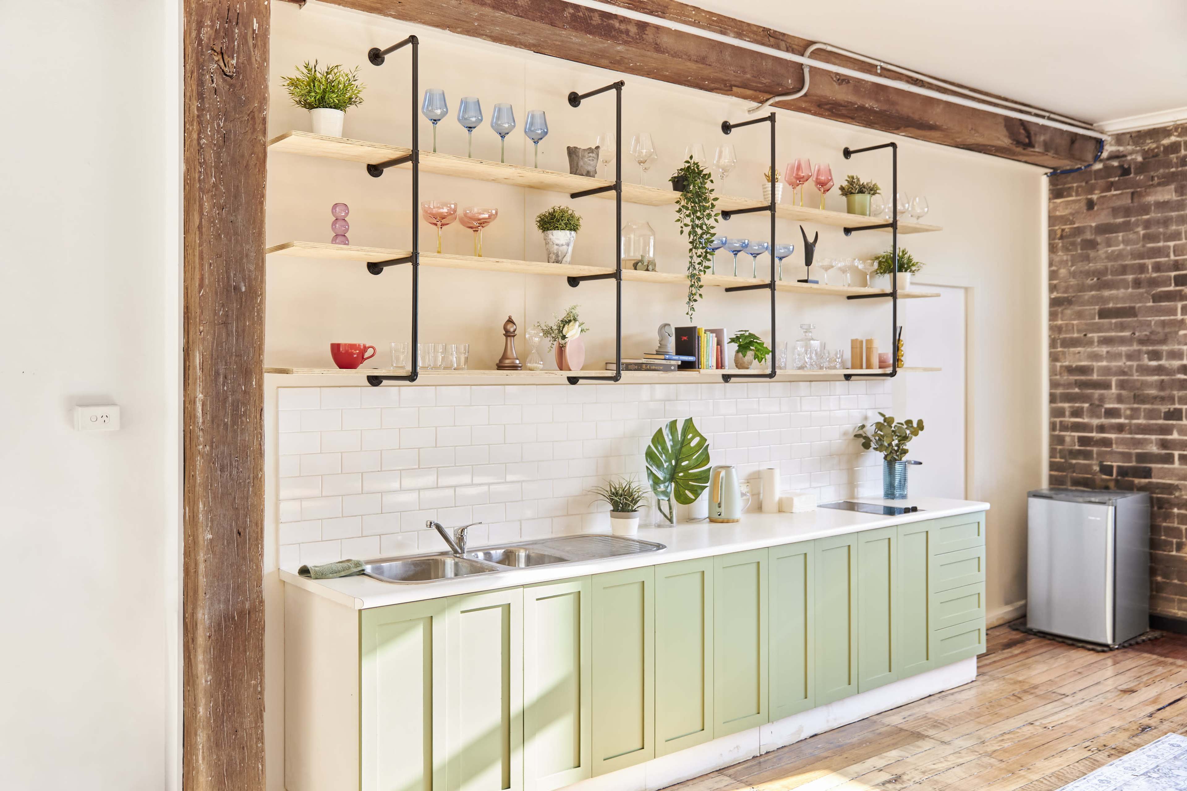 A kitchen with a green cabinetry base, a white tiled backsplash, and open shelving displaying various glassware and potted plants.