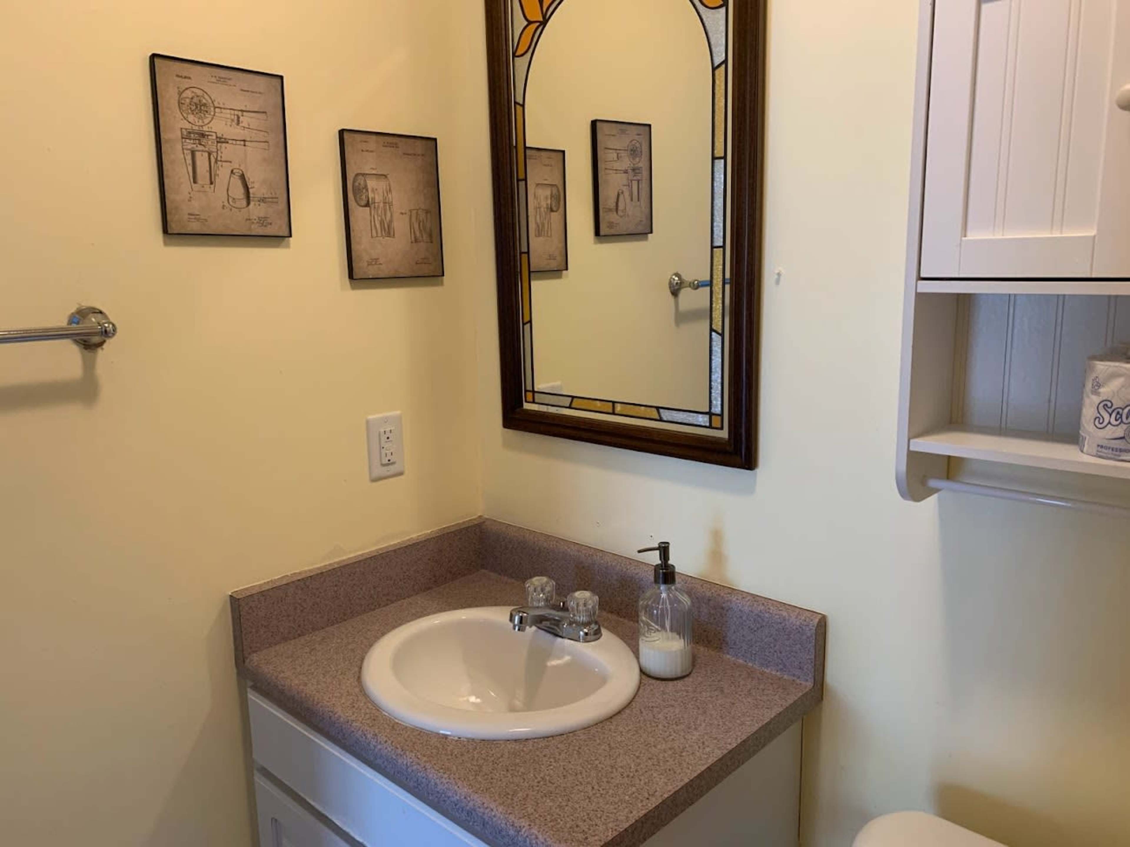 A small bathroom features a sink with a mirror above it, framed by decorative wall art and a storage shelf.