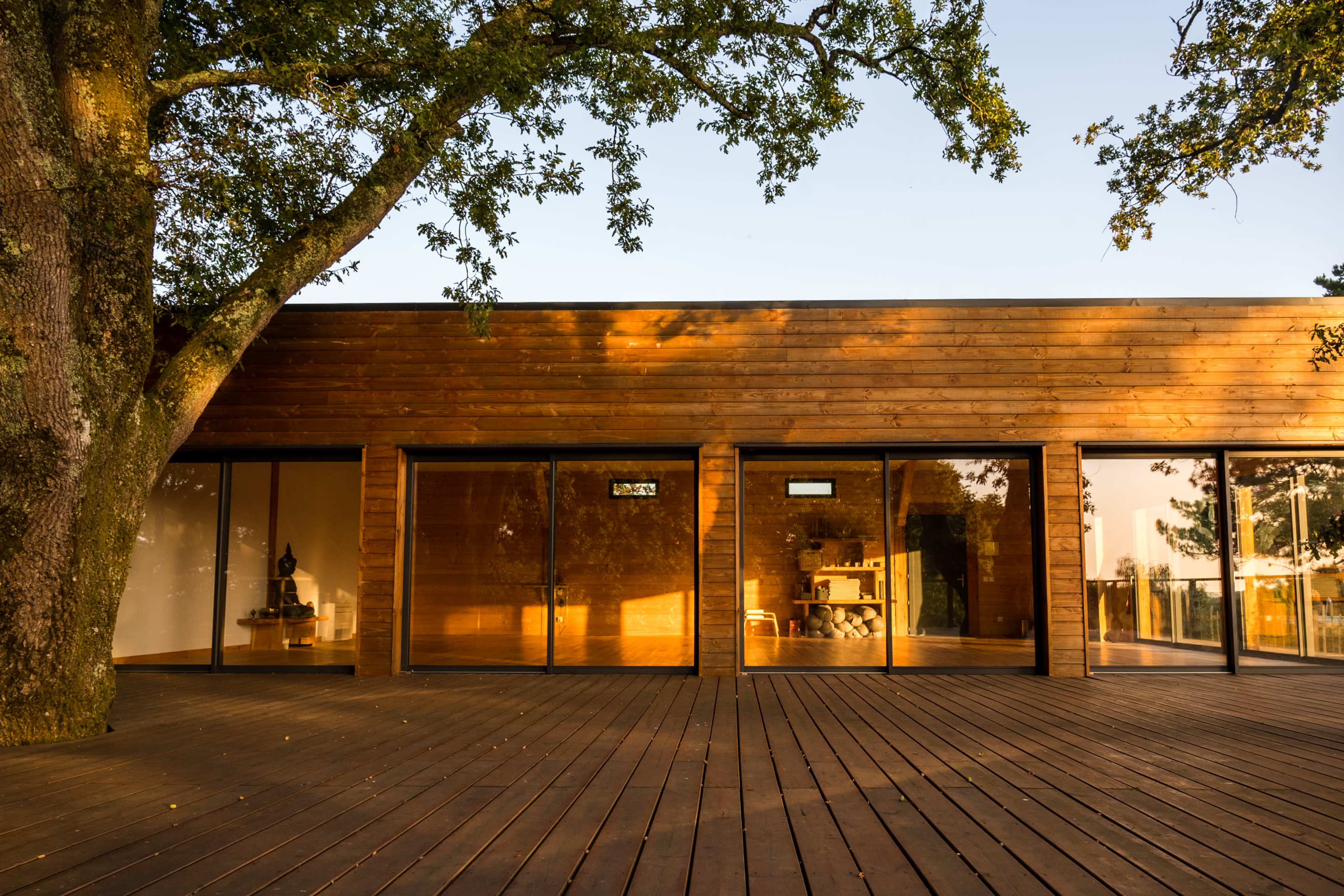 The image shows a wooden single-story house with large glass windows, surrounded by trees and a wooden deck.