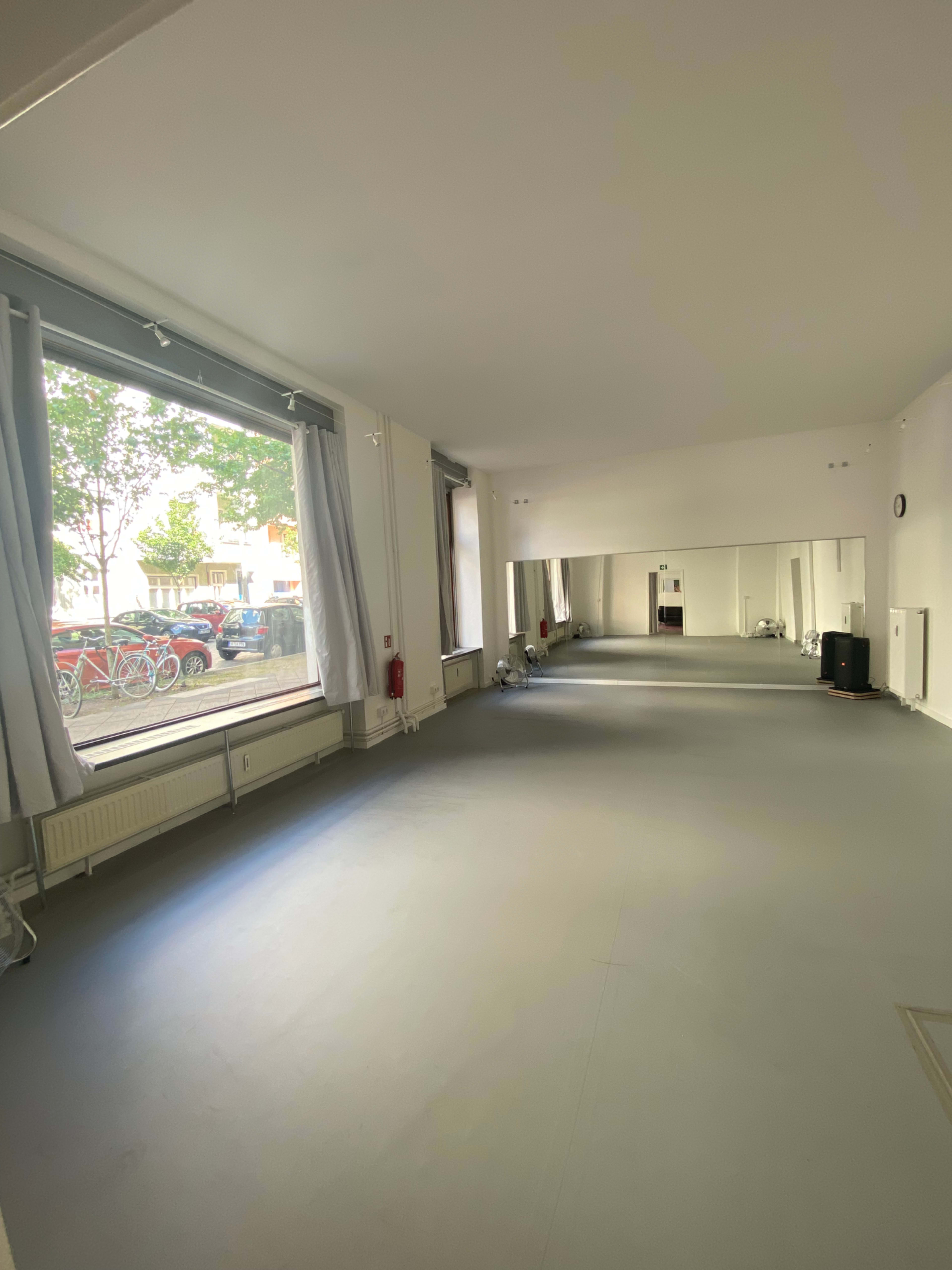 The image shows an empty room with large windows, a mirror on one wall, and smooth gray flooring.