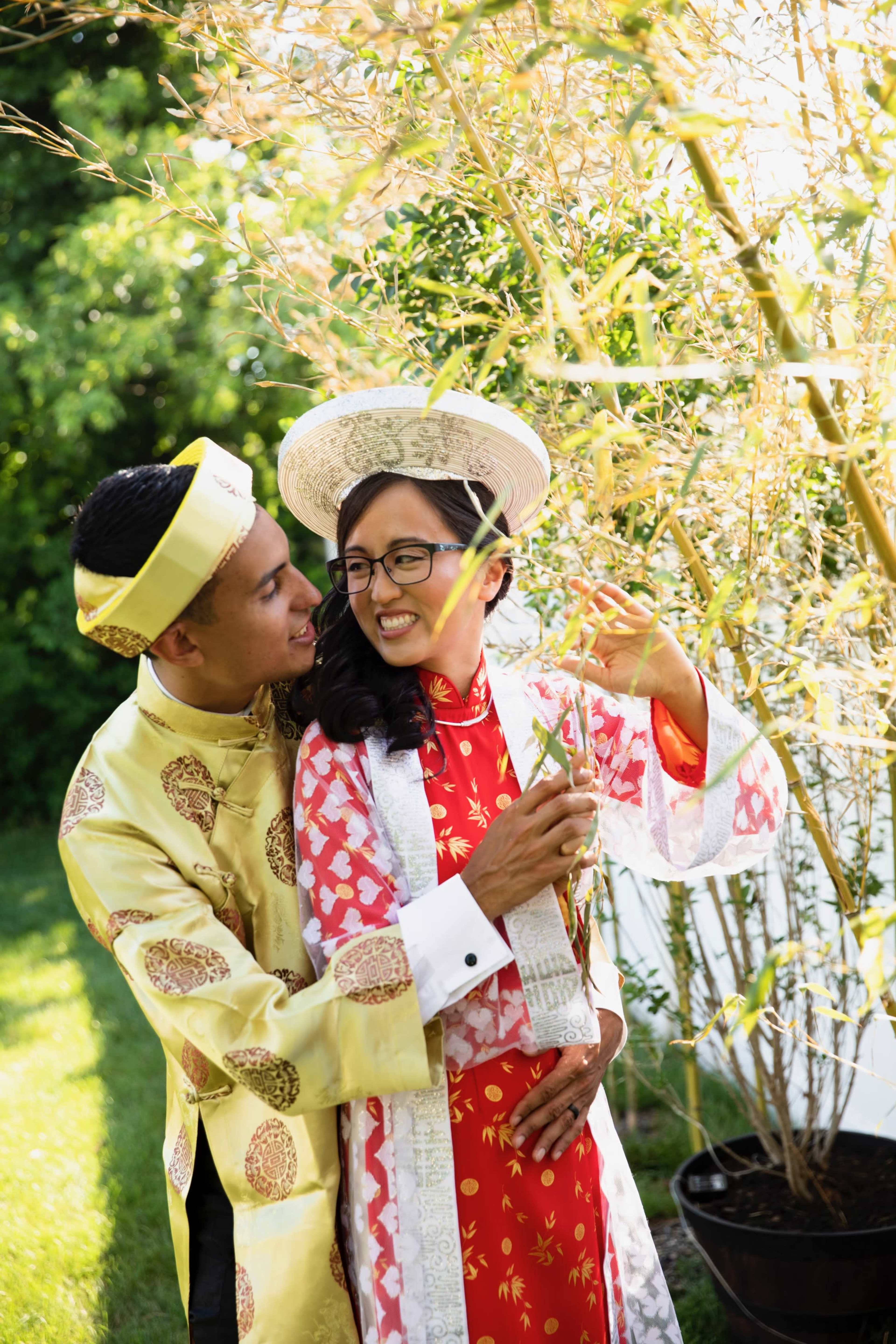 A couple dressed in traditional attire stands together in a garden, surrounded by bamboo plants.